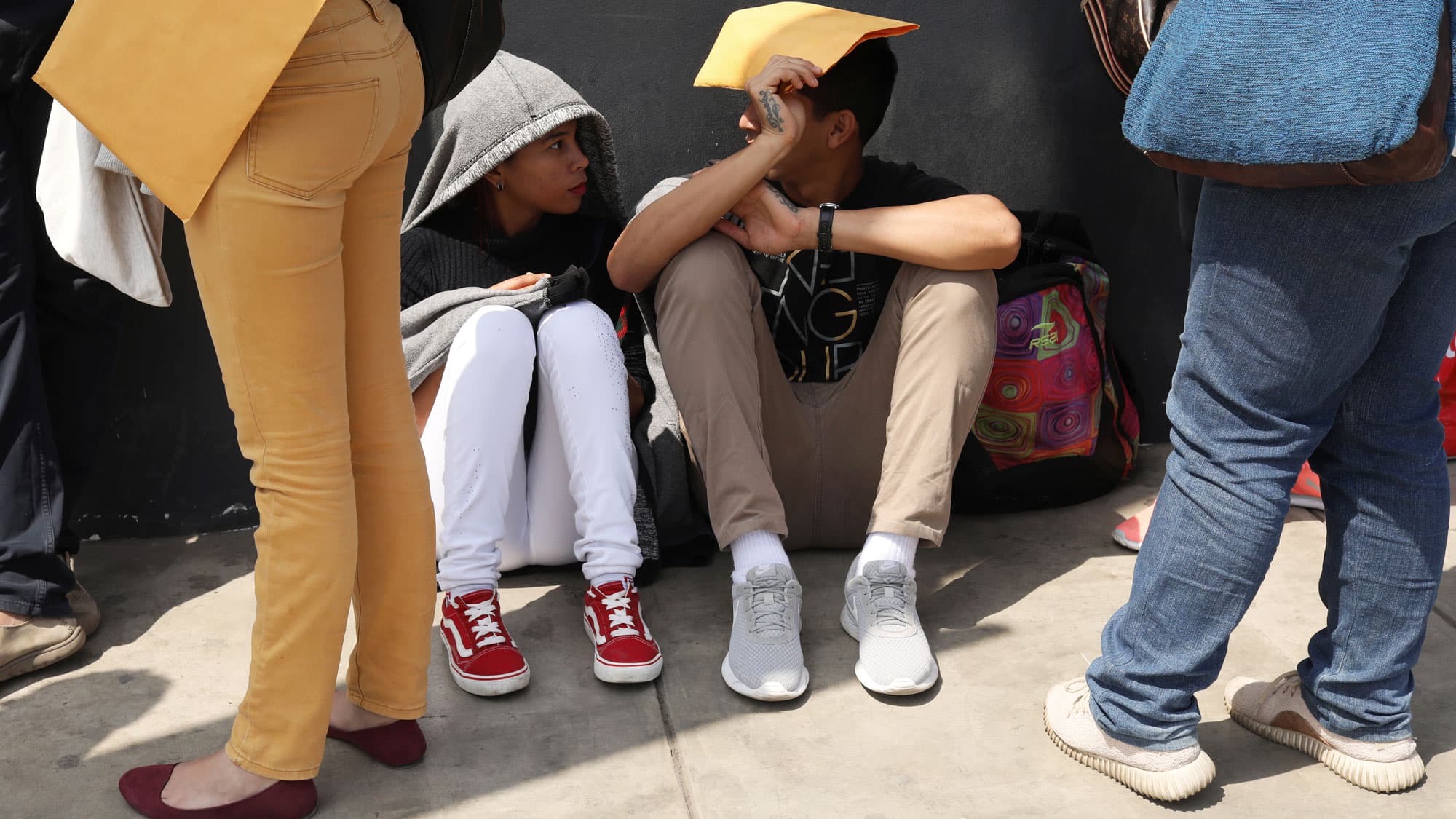Venezuelan migrants are shown standing and also sitting while they wait in line to get the needed paperwork for a temporary residency permit in Lima, Peru.