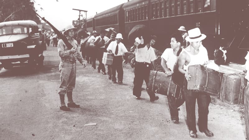 A solider with a rifle watches while men carrying suitcases line up next to a train.