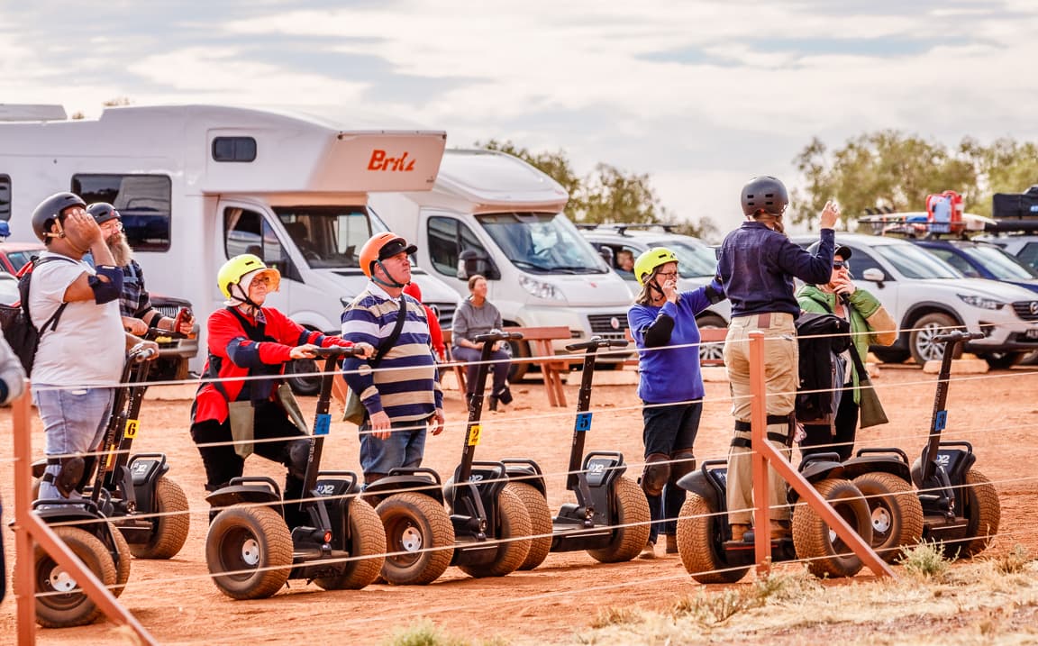 Tourists stand on segues wearing helmets looking off at Uluru as the sunset brings vast color changes and shadows to the face of the rock.