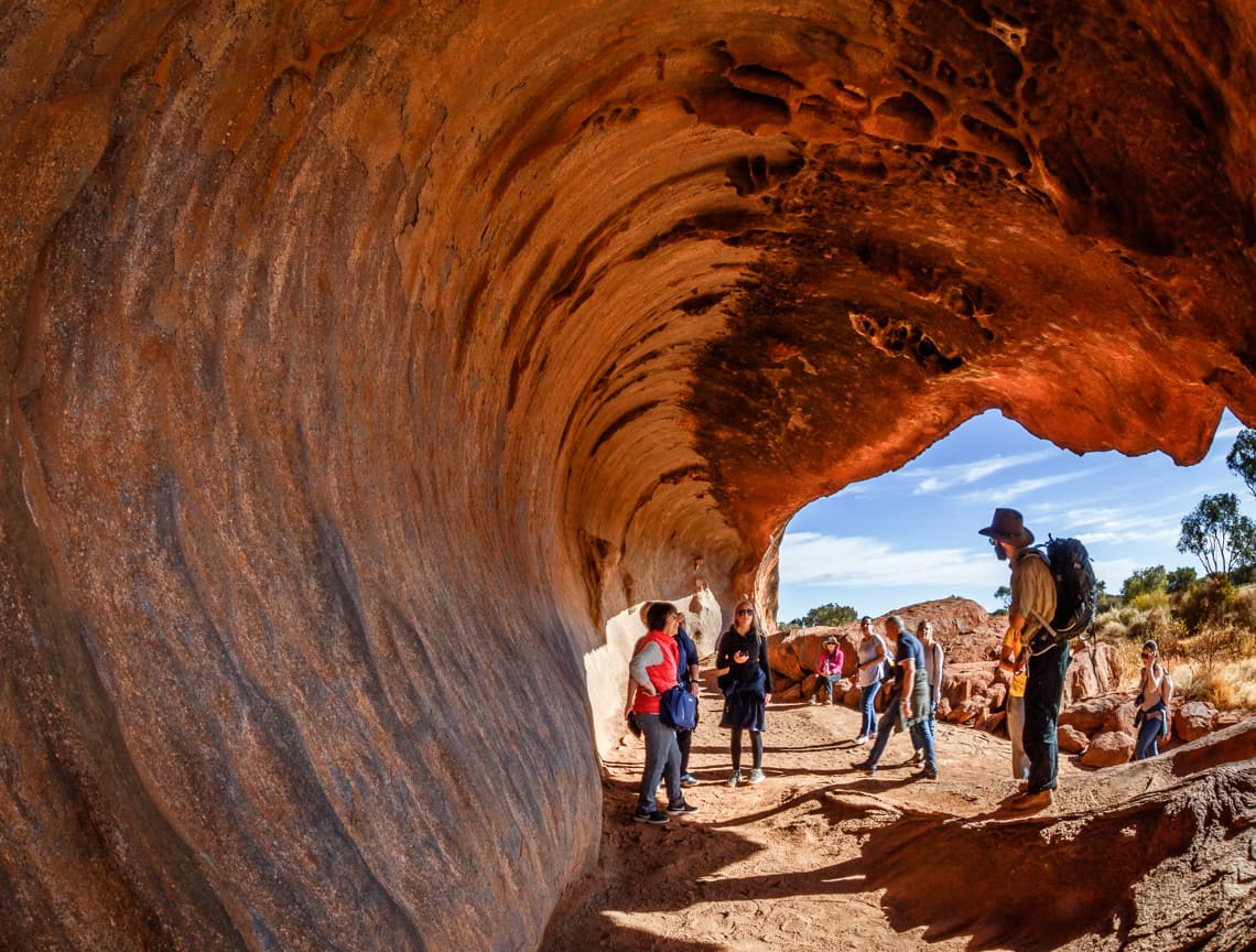 Guides stand under a rock overhang at the base of Uluru explaining the unique rock formations.