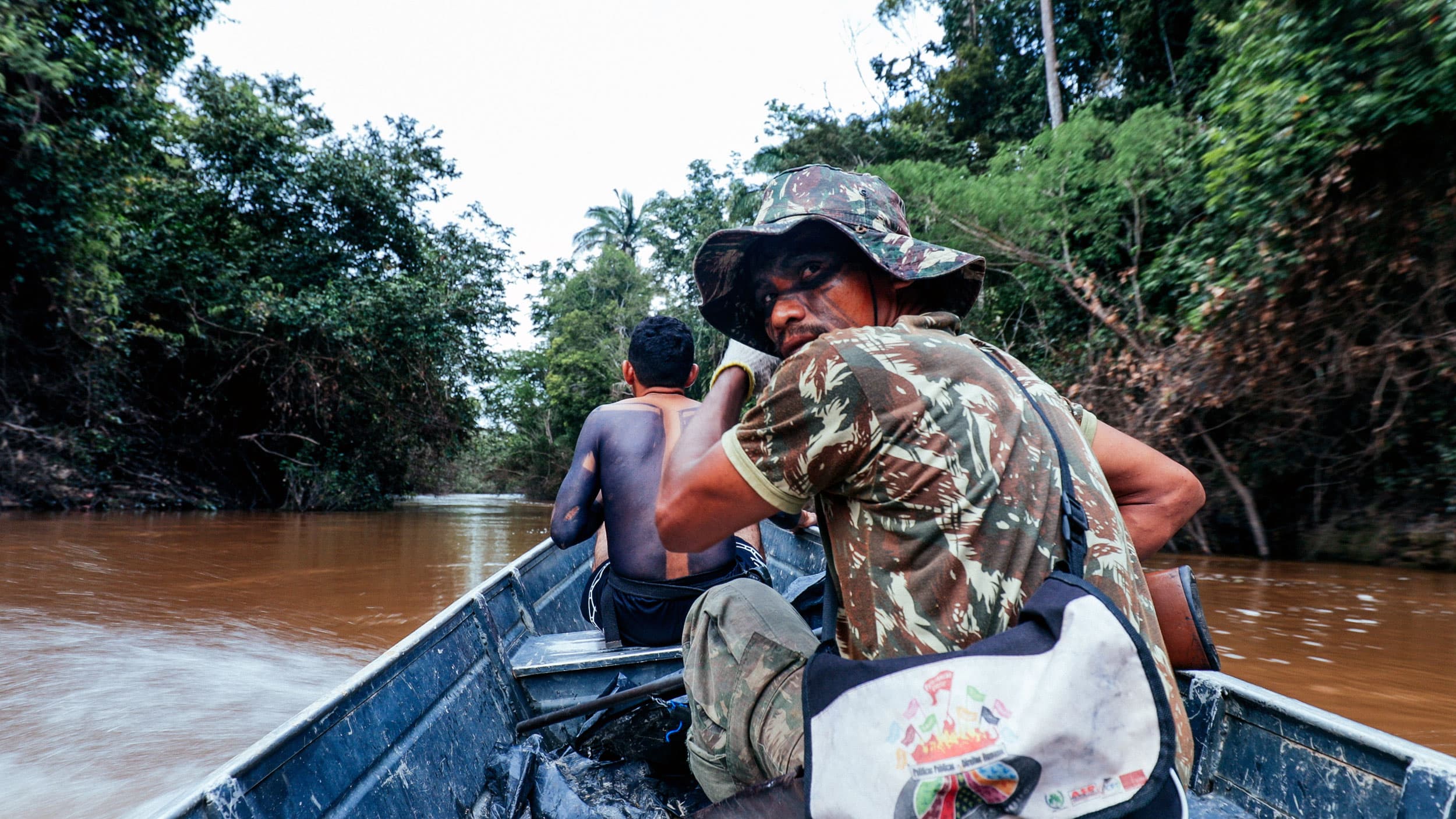 A man in camouflage points at something on the river bank. He holds a long rifle in his hand.