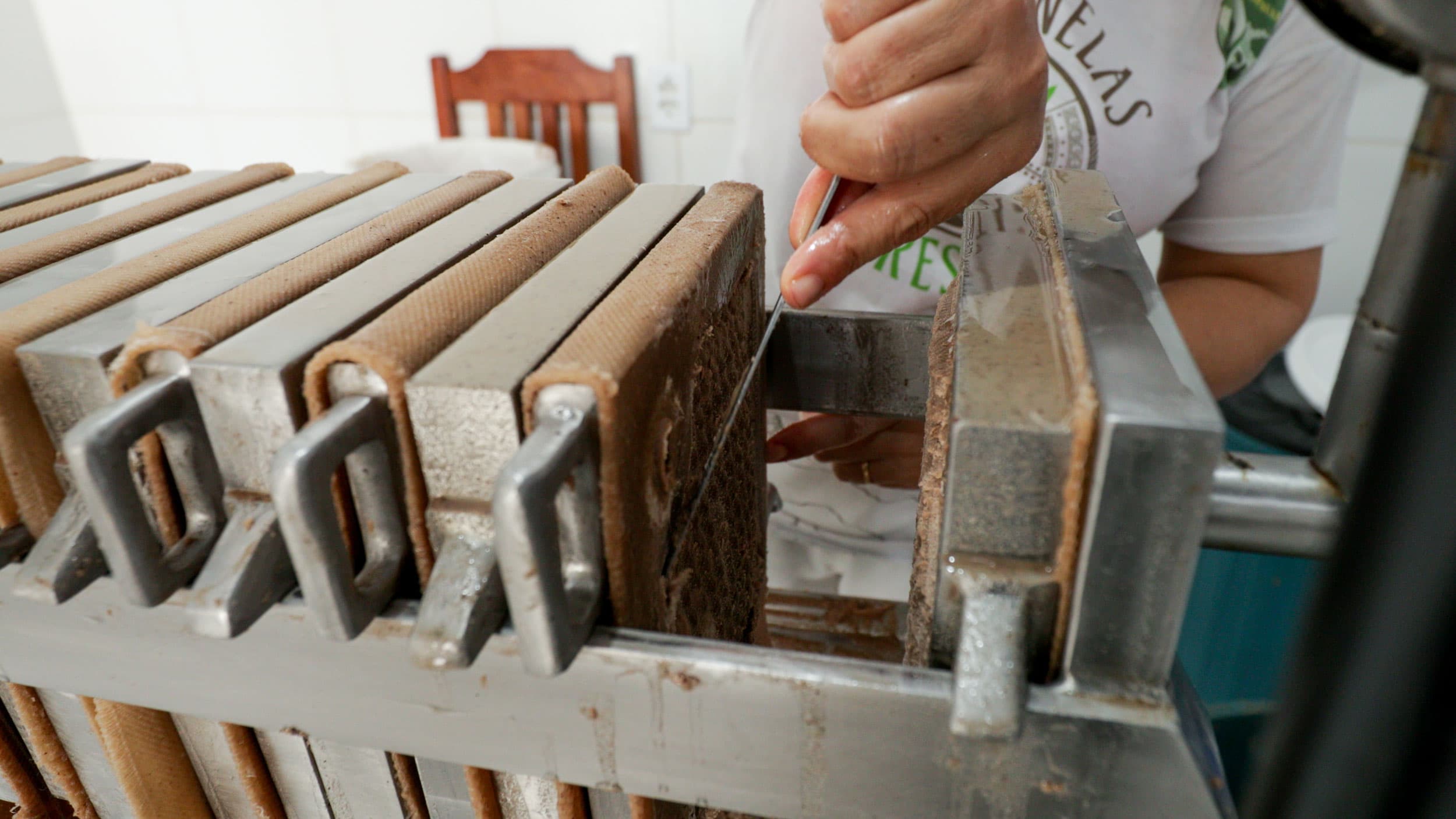 A brown paste oozes out of a press as a human hand uses a knife to scrape it