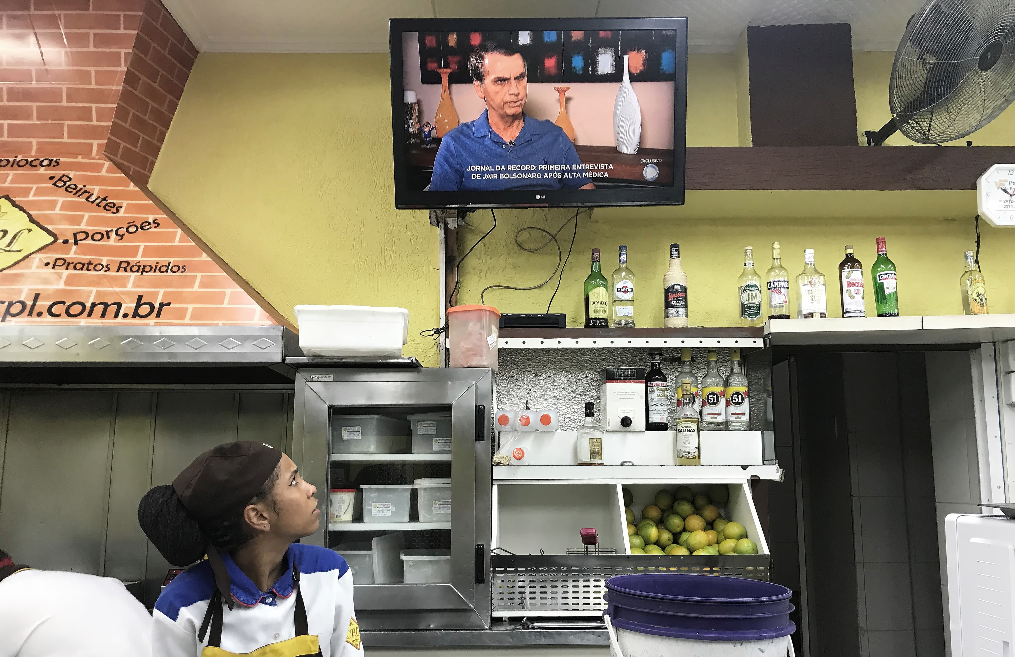 a woman working in a kitchen watches the tv over her shoulder