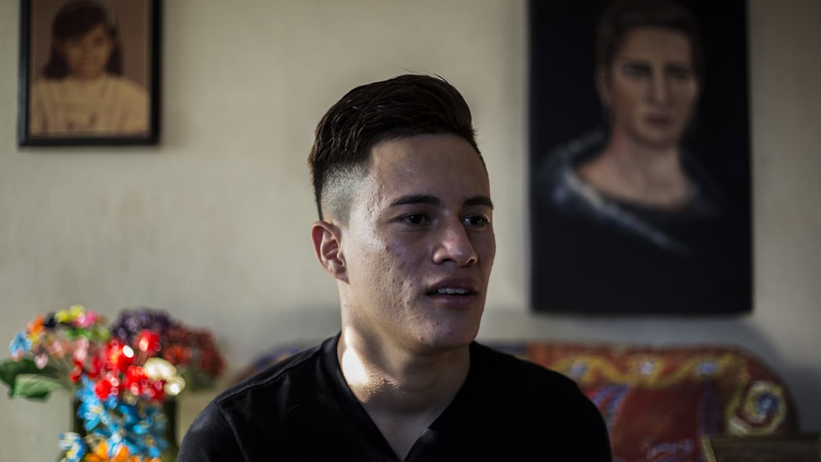 Daniel Alemán wears a black shirt in a room with two paintings on the wall and a blurry bouquet of flowers.
