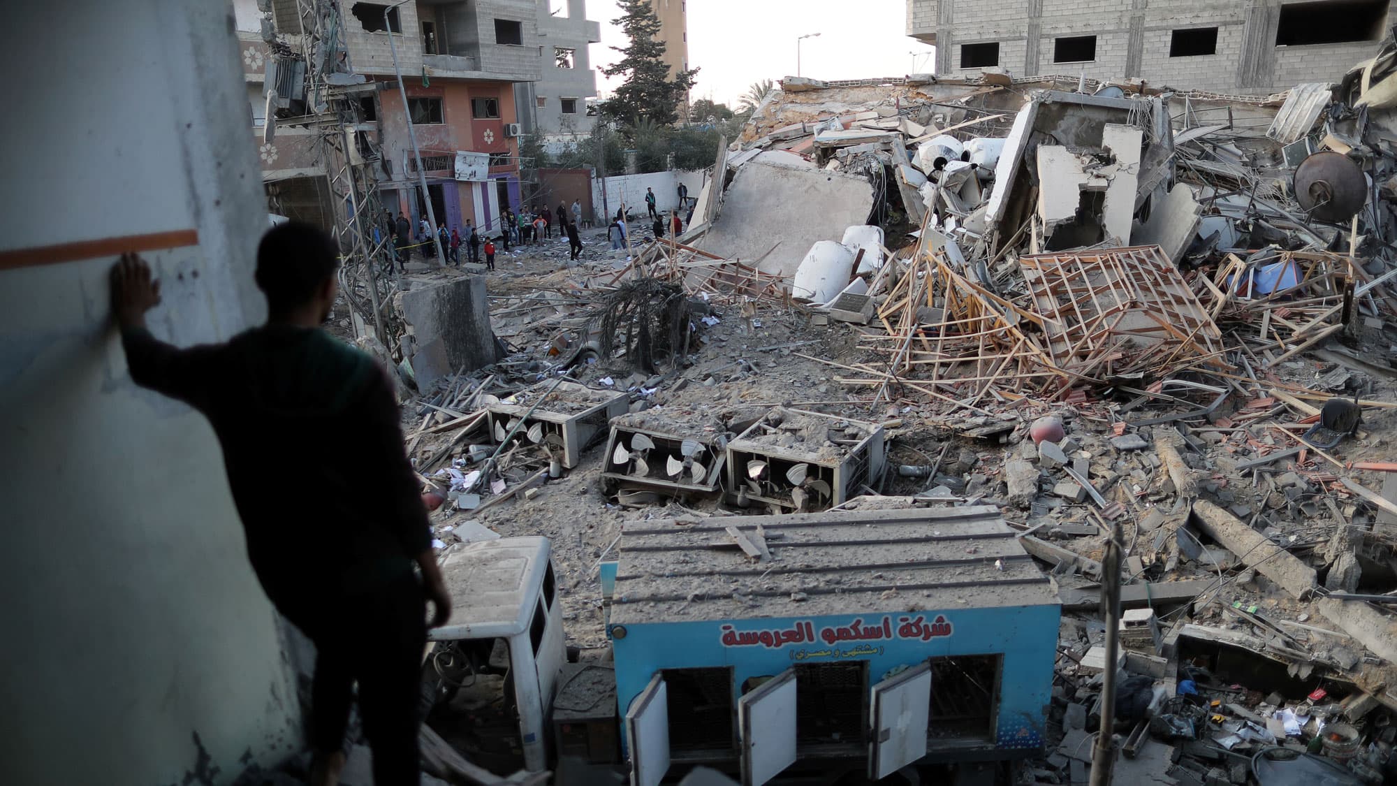 A Palestinian man is shown in shadow in the near ground looking out at the destroyed remains of Hamas's TV station building.