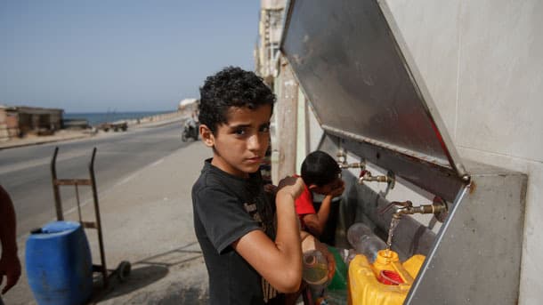 A boy with dark hair fill a jug of water at a tap provided by a mosque