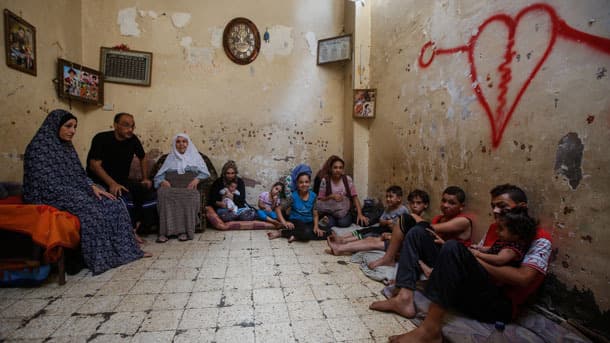 Fifteen family members in Gaza sit in the living room with a red heart spray painted on the cream-colored wall.