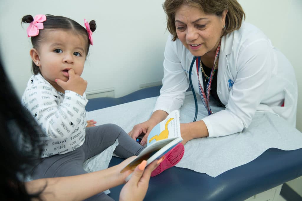 A toddler sits on the examination table. A doctor is to her right