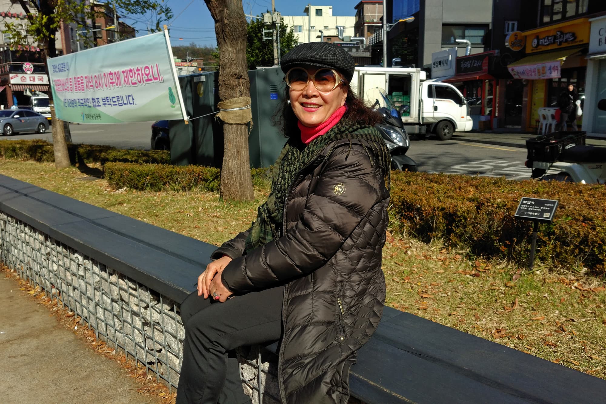 Woman in black coat and hat poses for camera on street ledge.