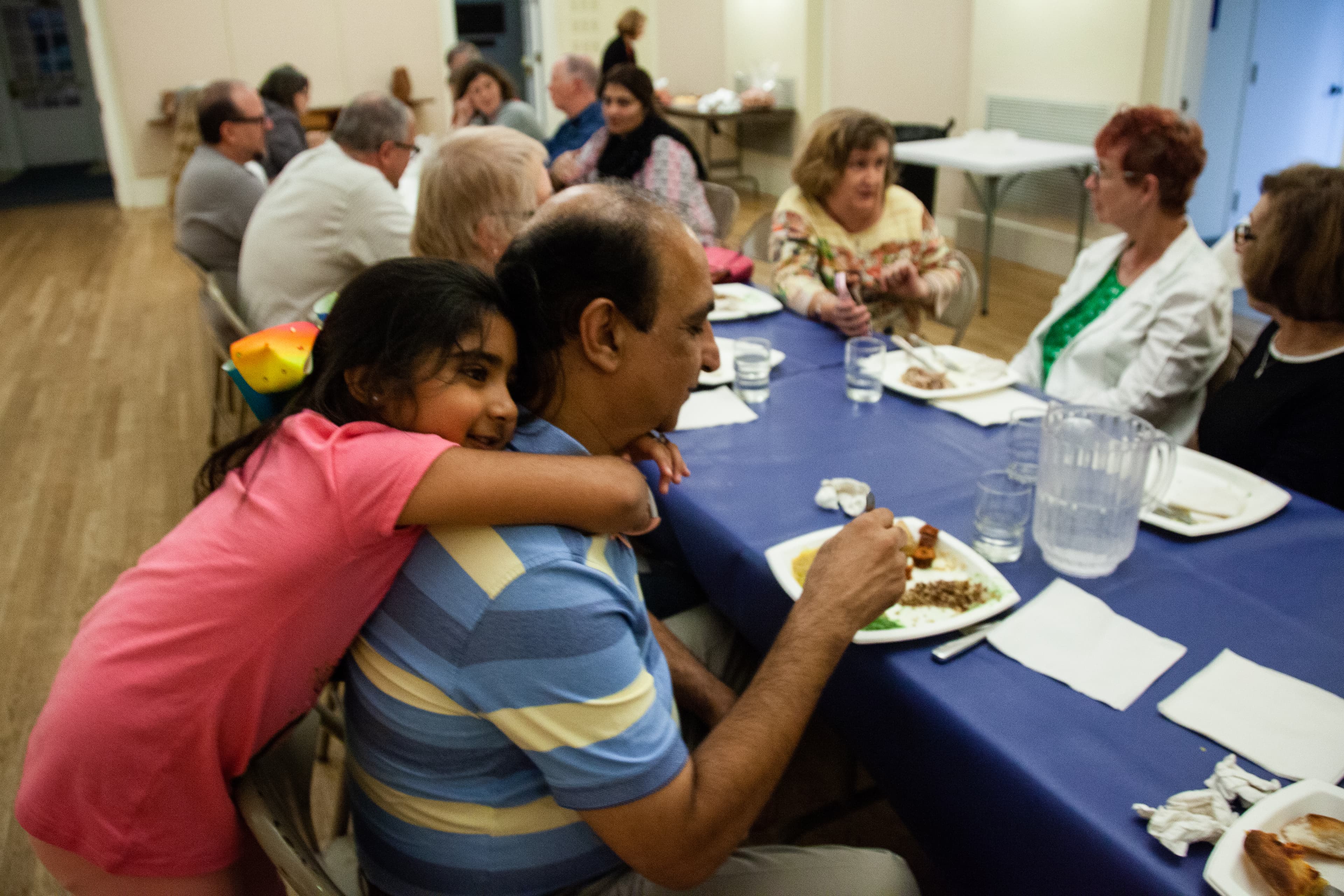 young girls hands onto back of man by neck, who is sitting at long communal table that is covered in blue, plastic tablecloth