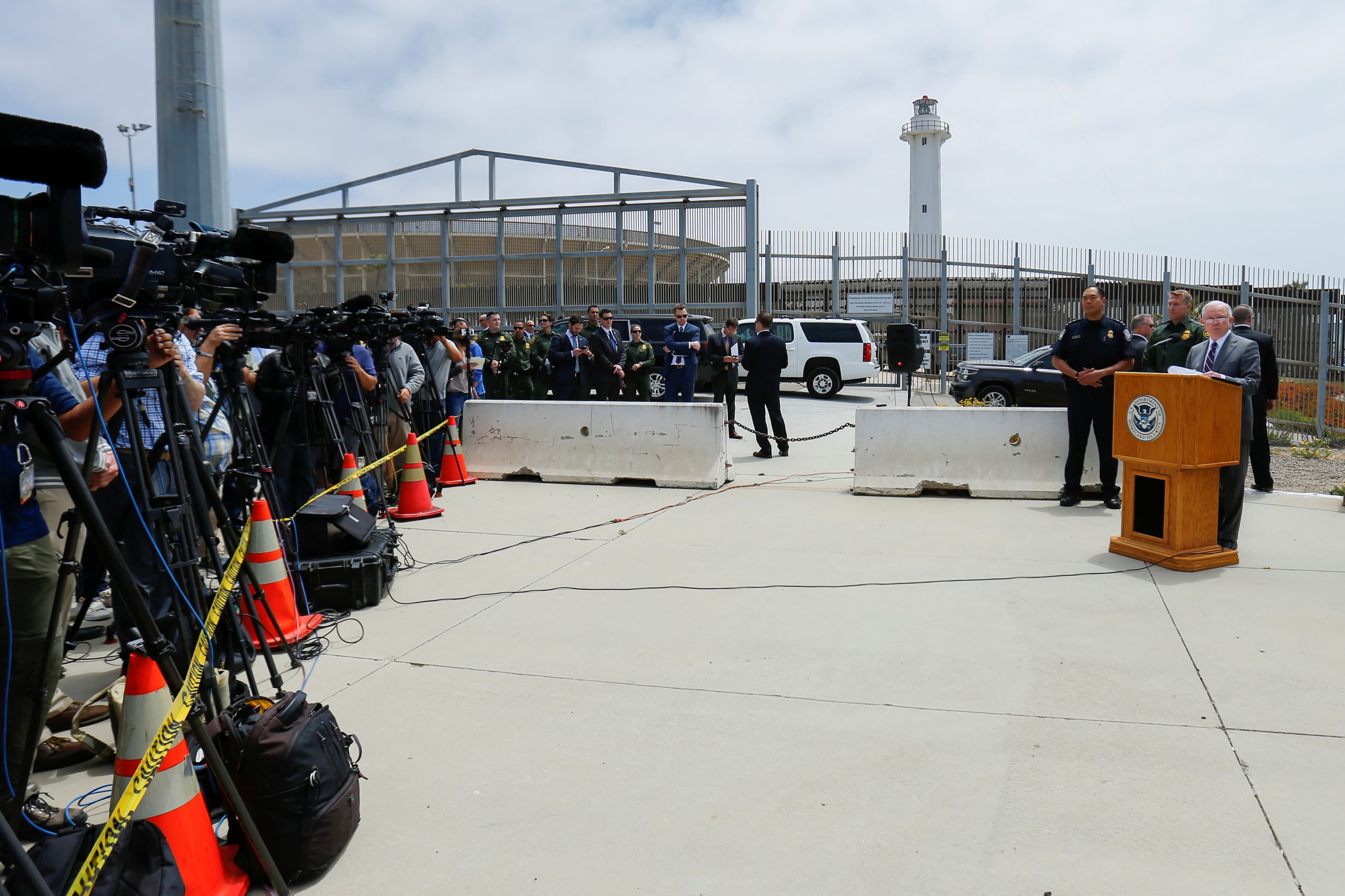 Man standing at podium, speaking, with fence behind him and people holding cameras in front