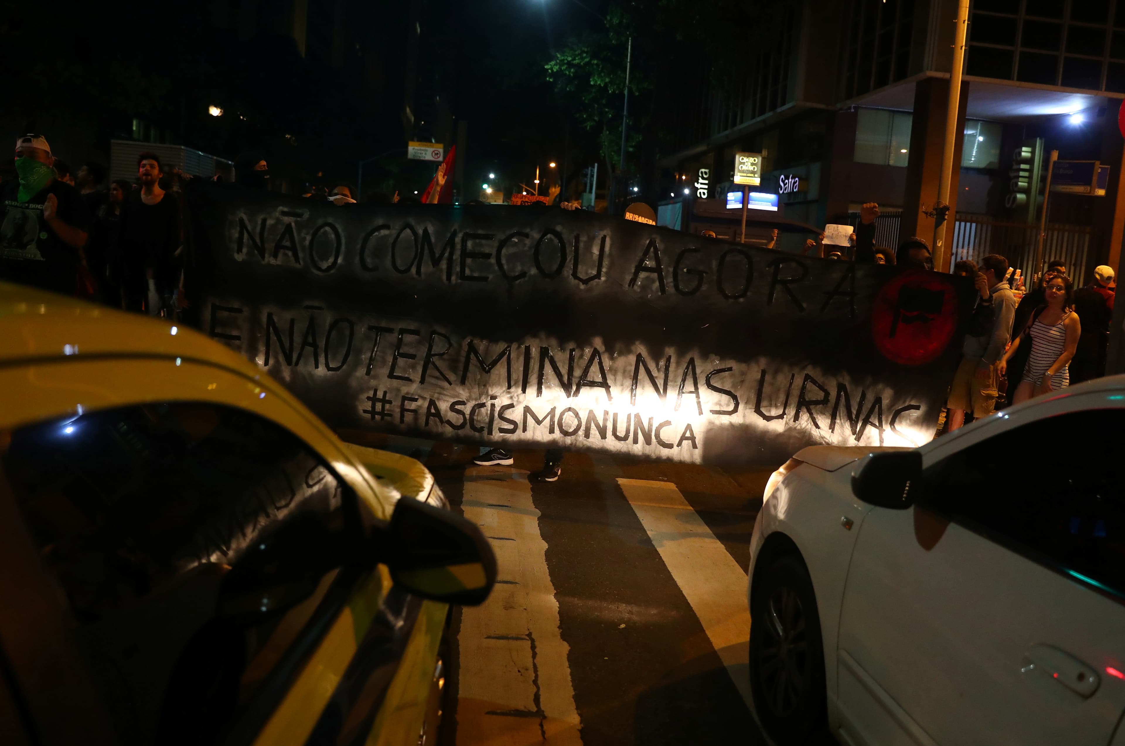 two cars flash their lights on a sign held by anti-Bolsonaro protesters