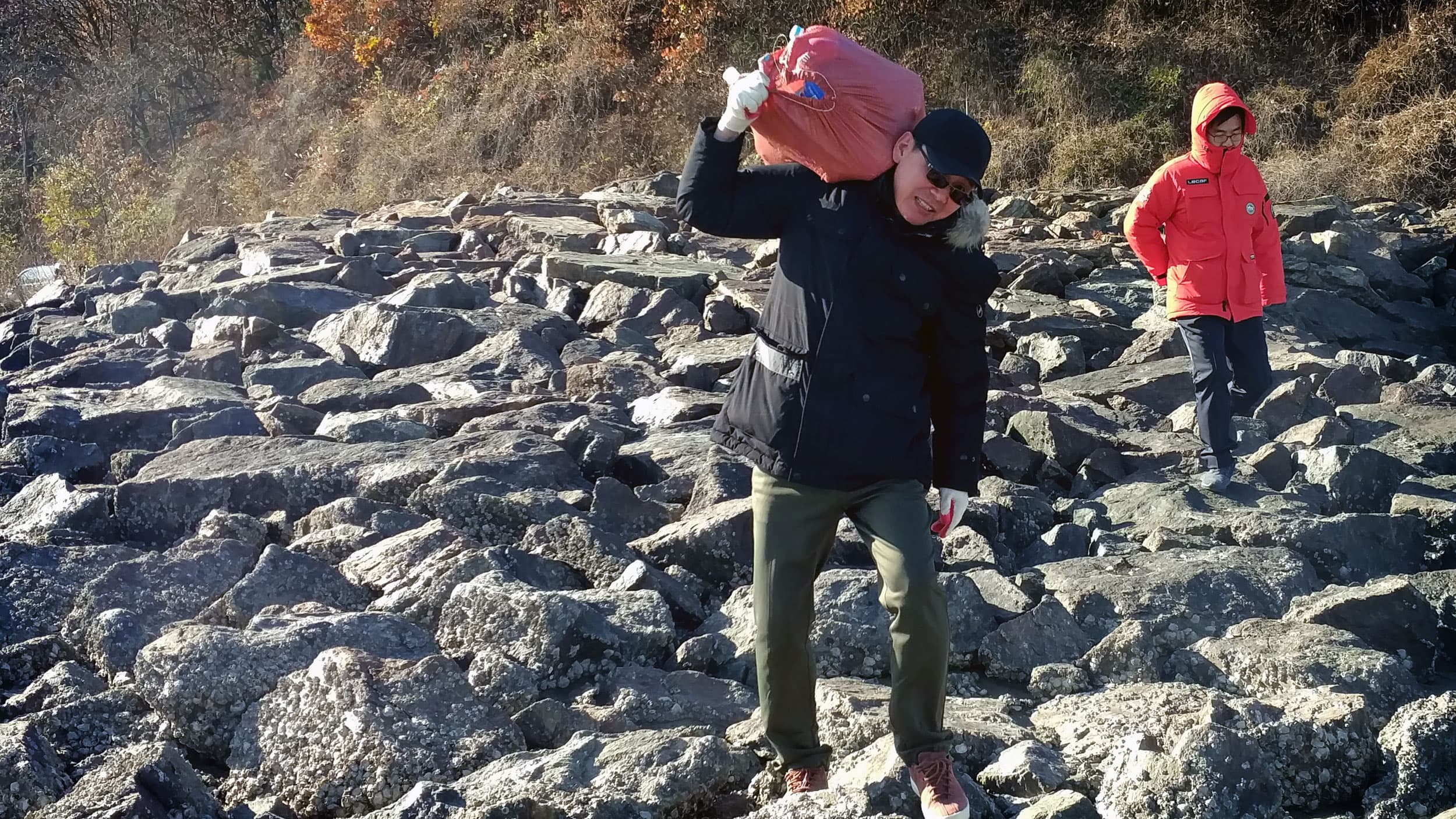 A man carries a brightly colored bag on his shoulders over rocky ground.