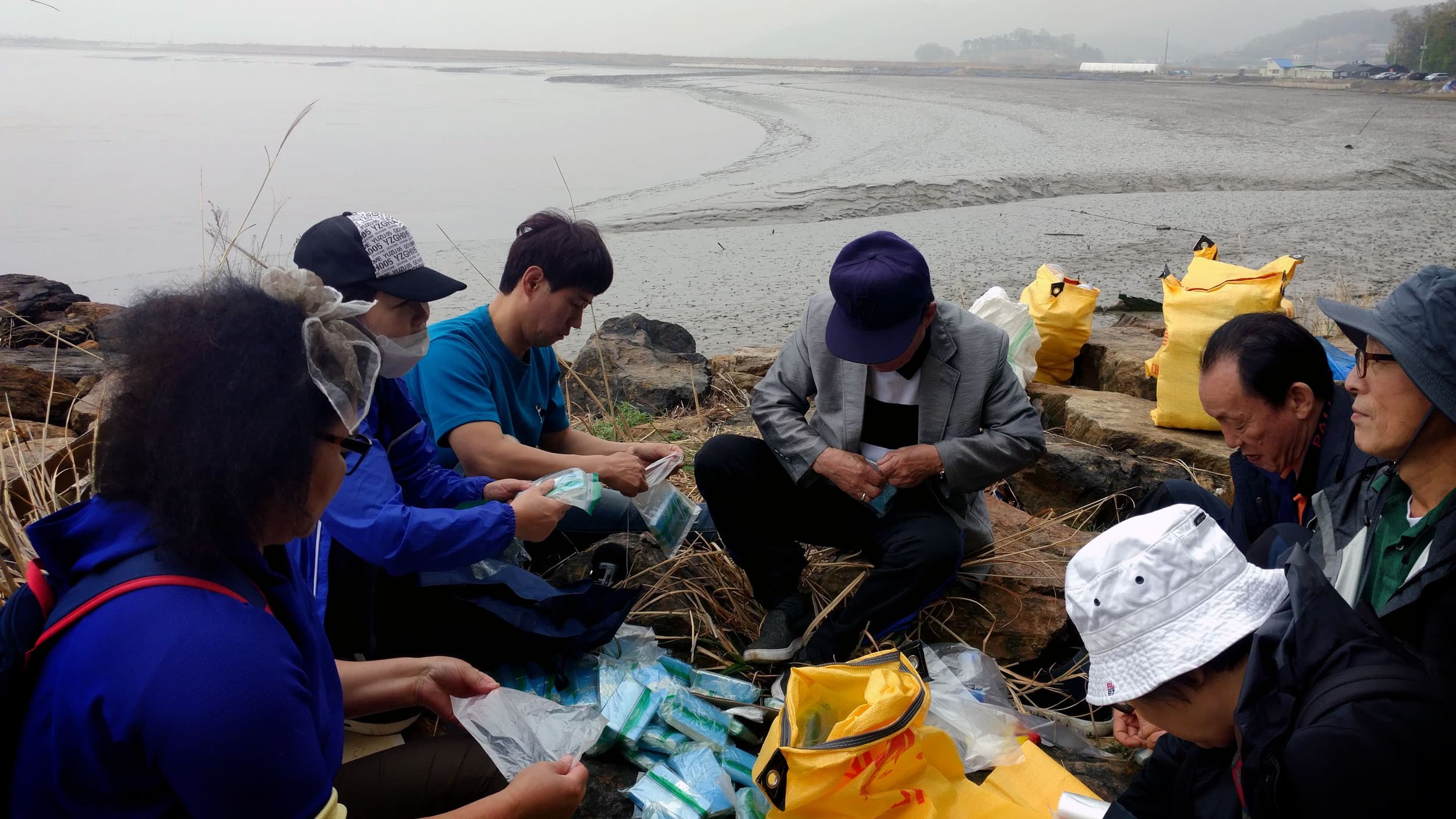 A group of people sit in a circle. Between them are blue bags and plastic bottles, which they are filling with rice, USB drives and tiny Bibles.