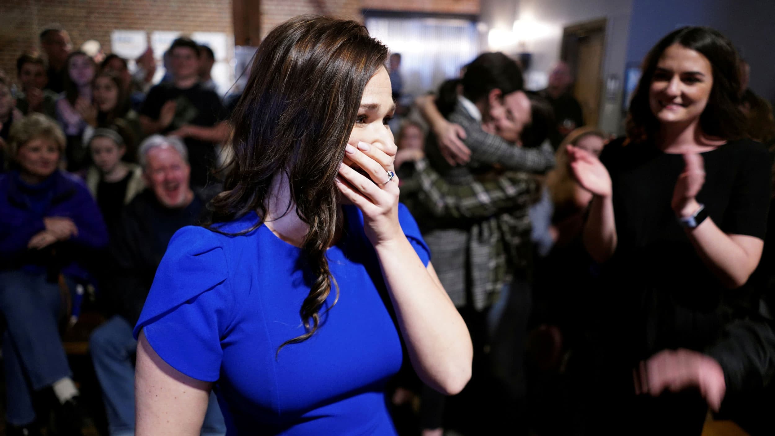 A woman in a bright blue dress covers her mouth with her hand in an emotional moment
