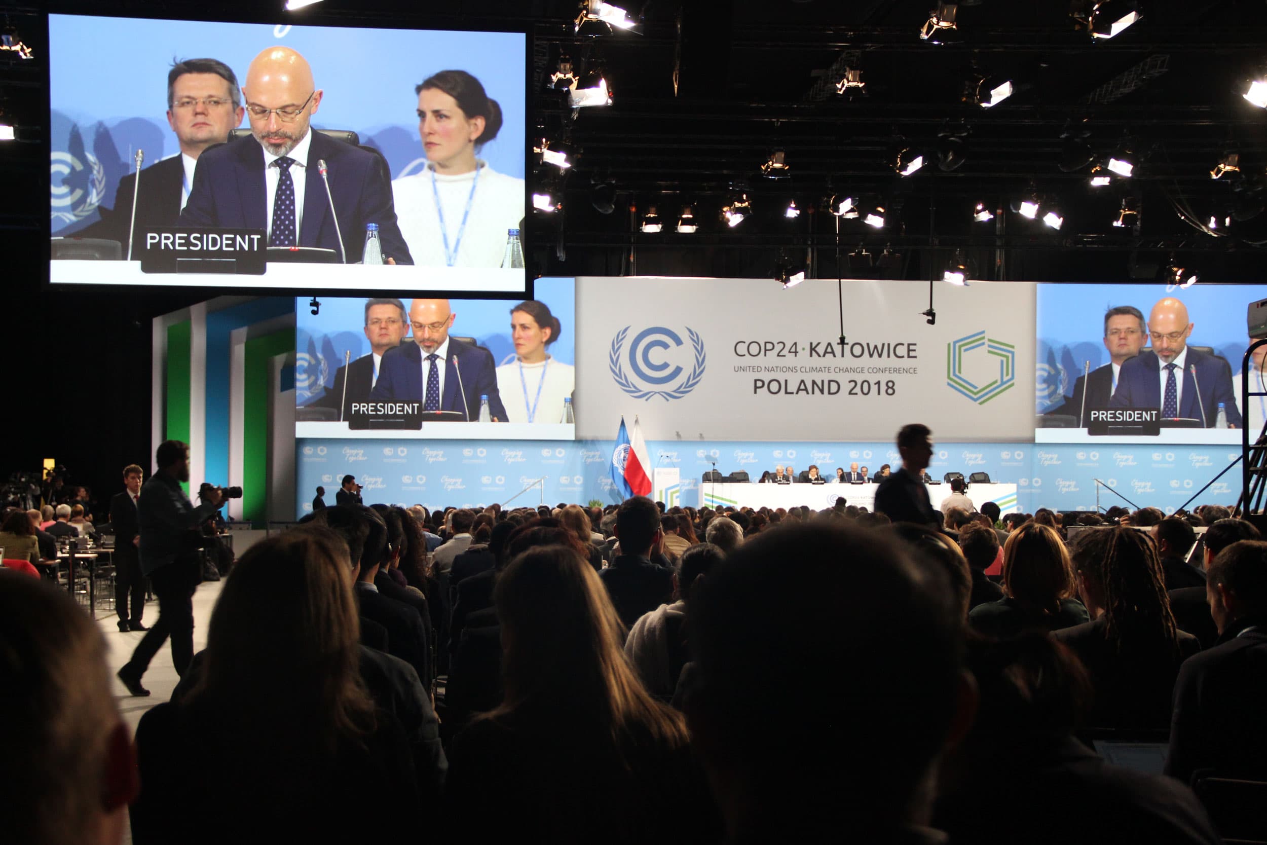 A crowd in a dark conference room watches a screen with a man that has a gavel and a sign that says 