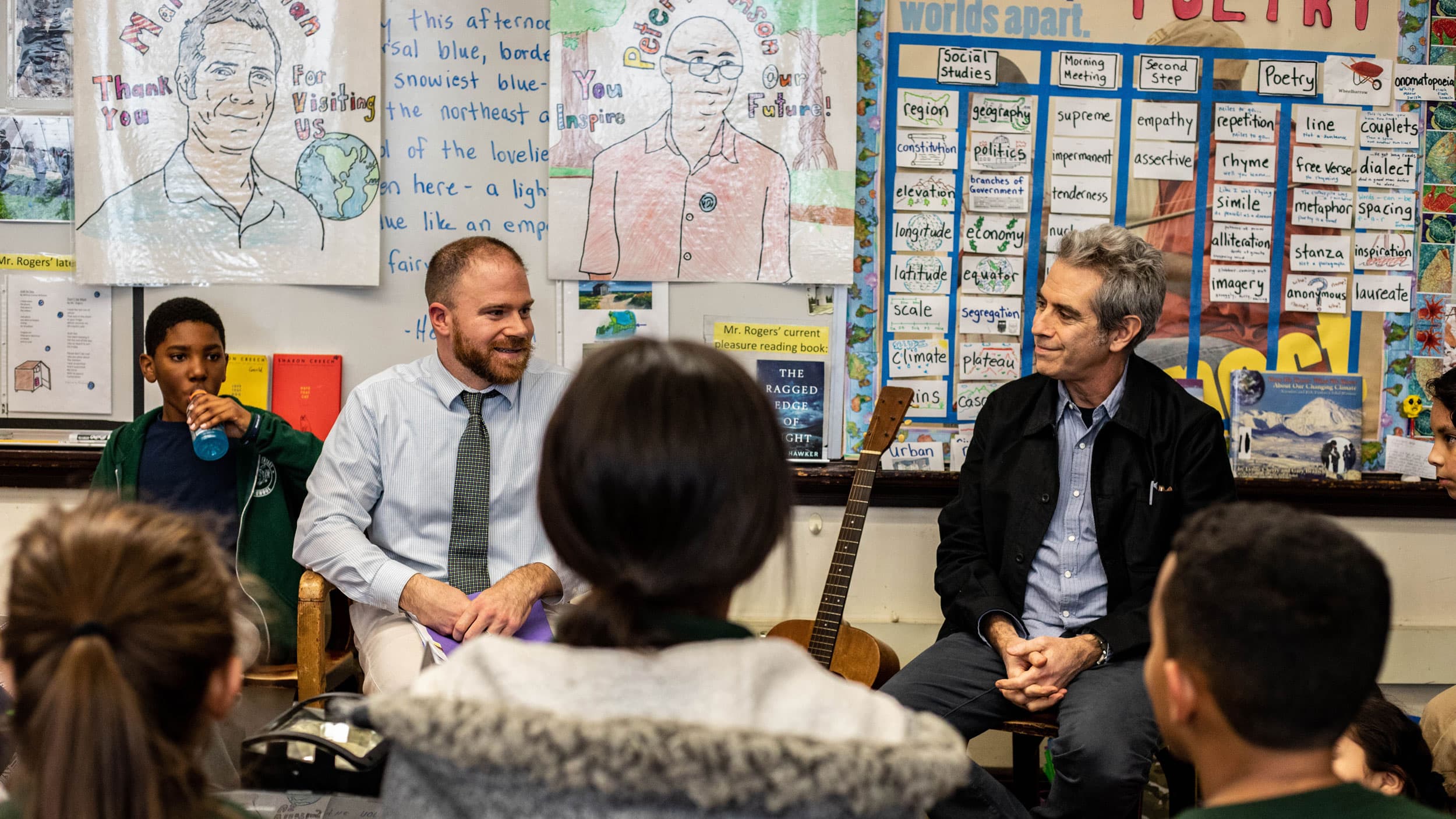 Two adults sit at the front of an elementary school classroom as children's heads fill the bottom of the frame.