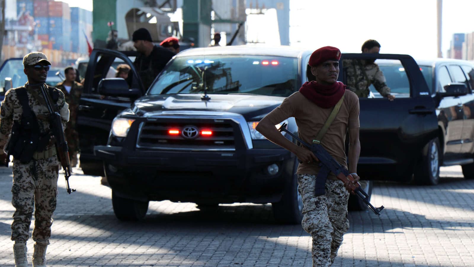 Two soldiers carrying guns stand in front of a black car.