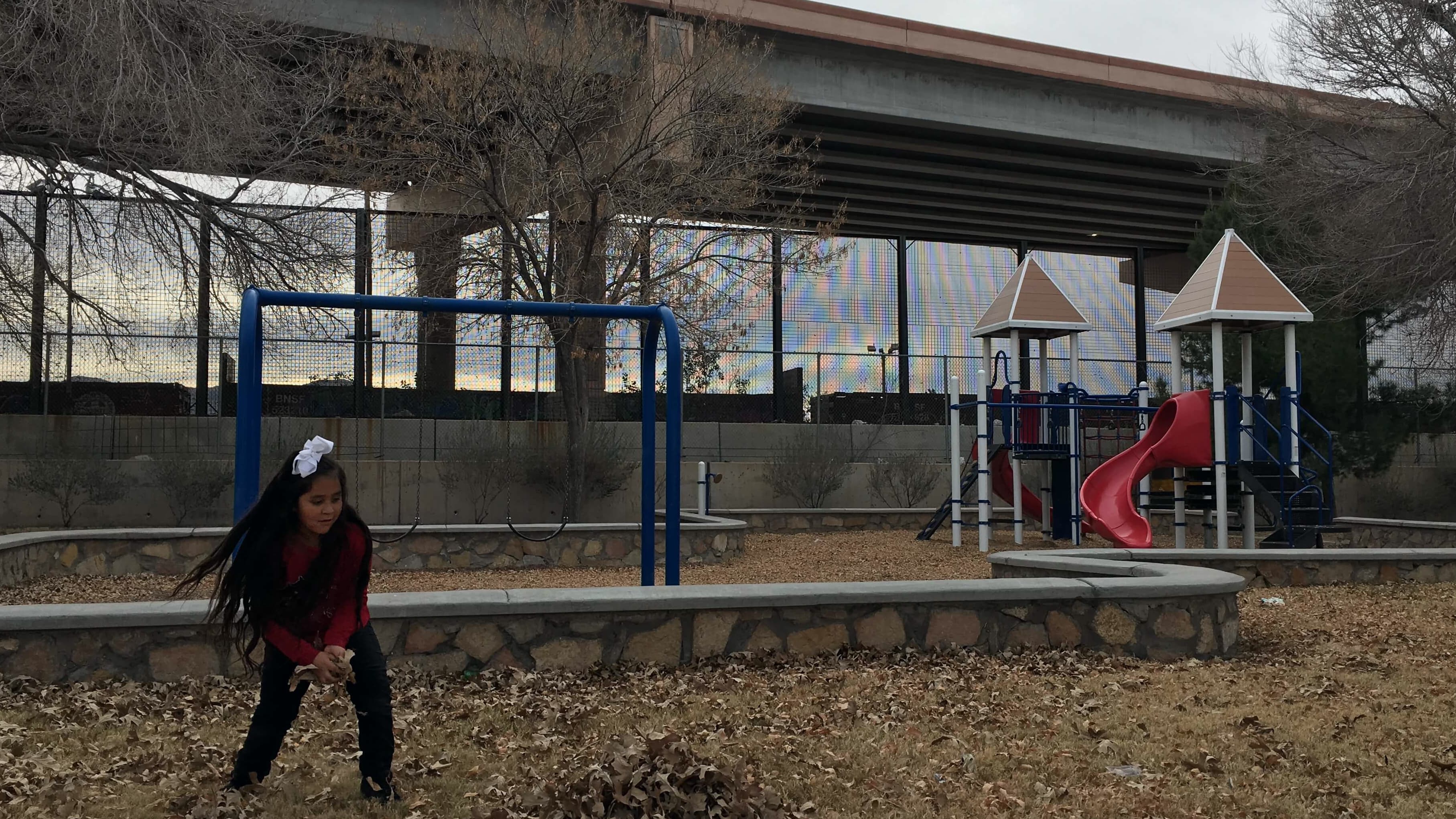 A little girl plays in a park with 18-foot steel border fence in backdrop.