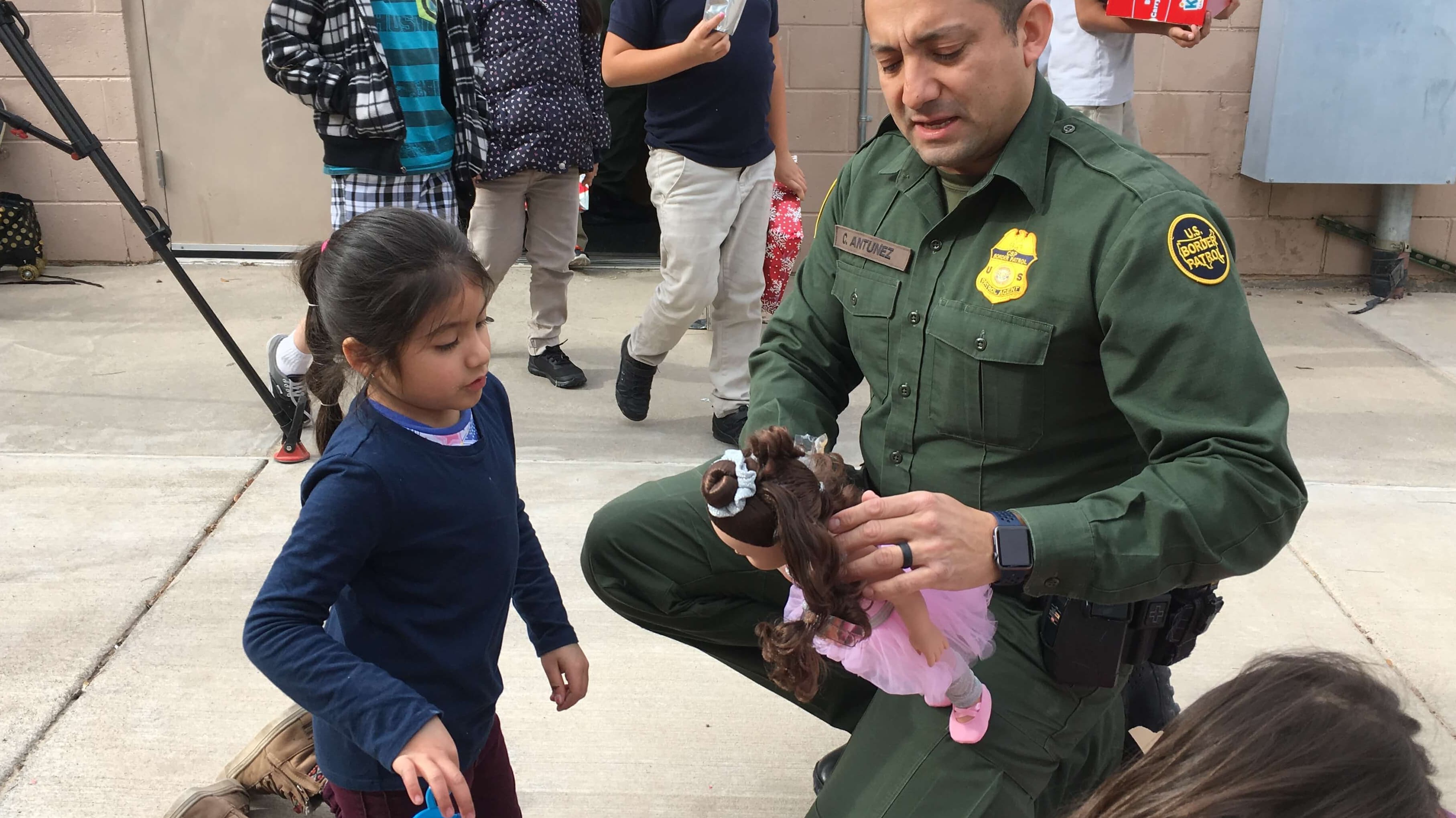 Border patrol agent helps a young girl dress a doll.