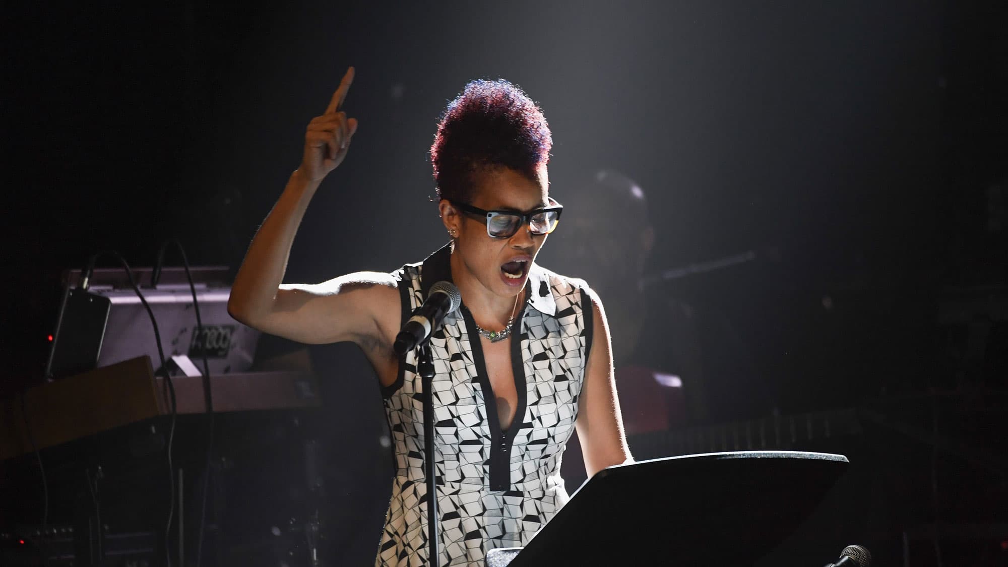 Woman with short hair wearing a black and white top raises her right arm while reading in front of a microphone.