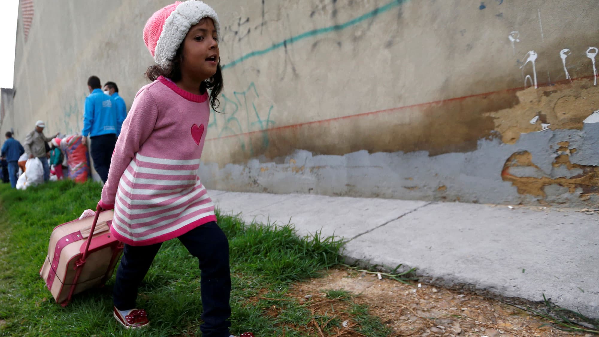 A Venezuelan migrant girl wearing a pink sweater with a heart on it, is shown heading to the exit of a makeshift camp with her belongings in Bogota, Colombia.