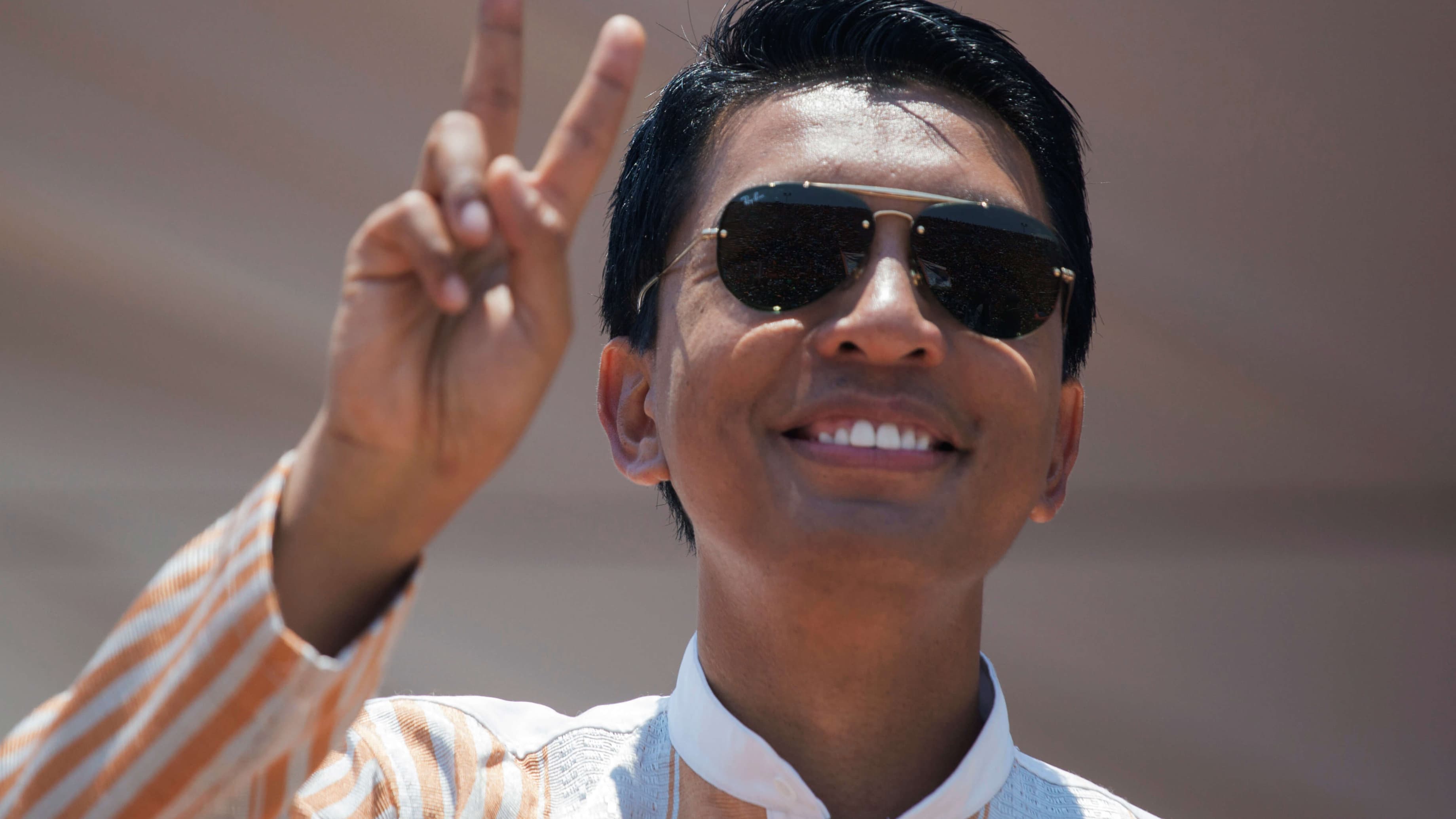 A man with glistening black hair wearing sunglasses waves to the crowd.