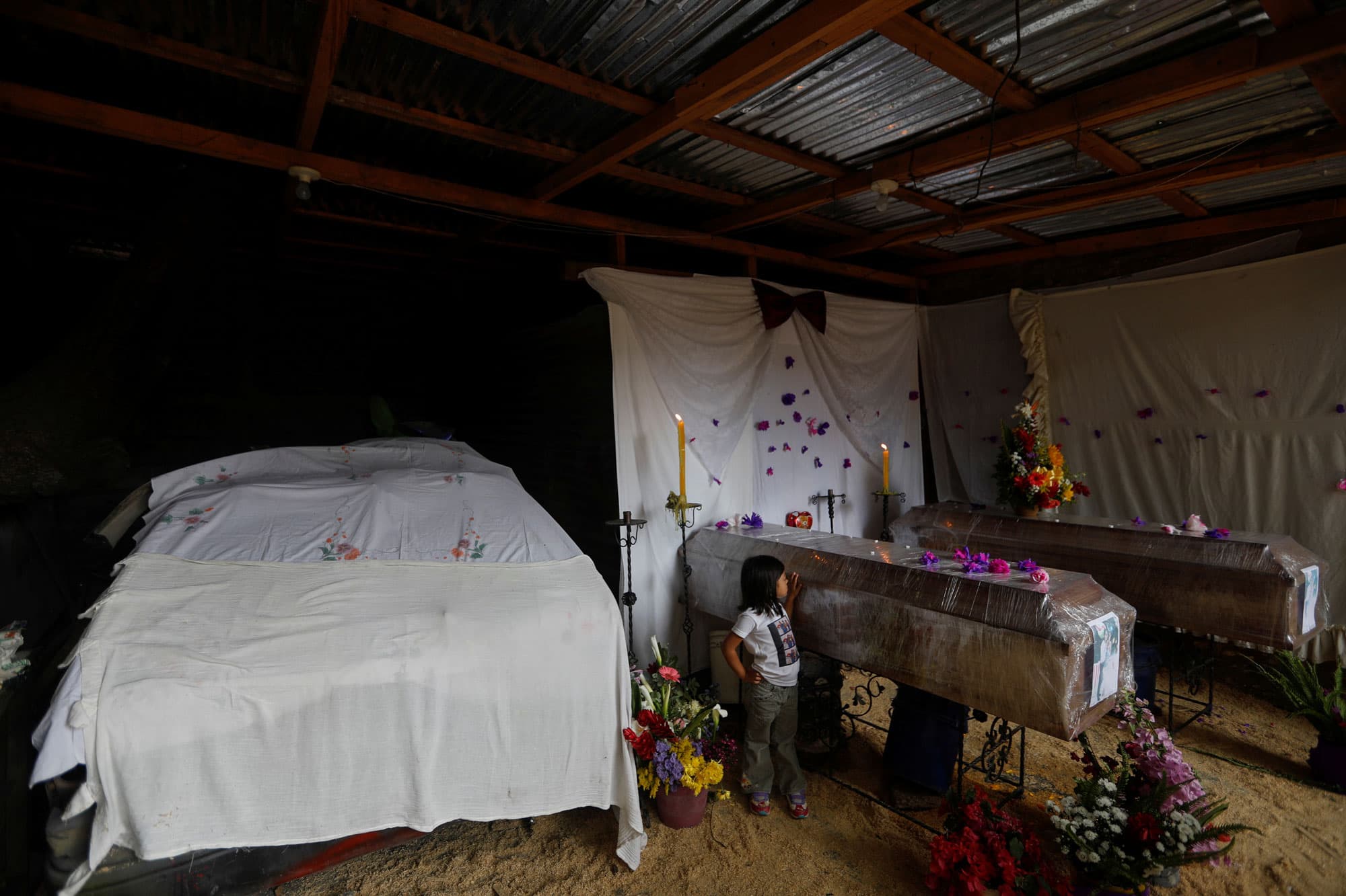 A young girl is shown standing next to two wooden coffins wrapped in plastic wrap.