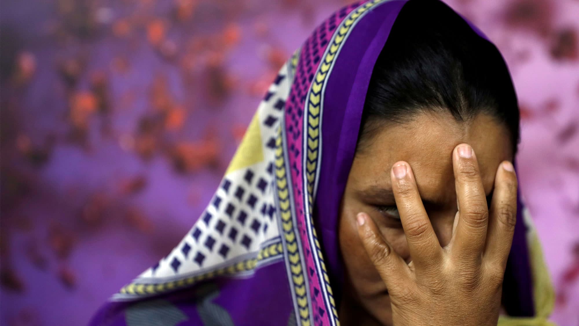 A woman is shown with a colorful purple and white scarf over her head and her hand held against her face.