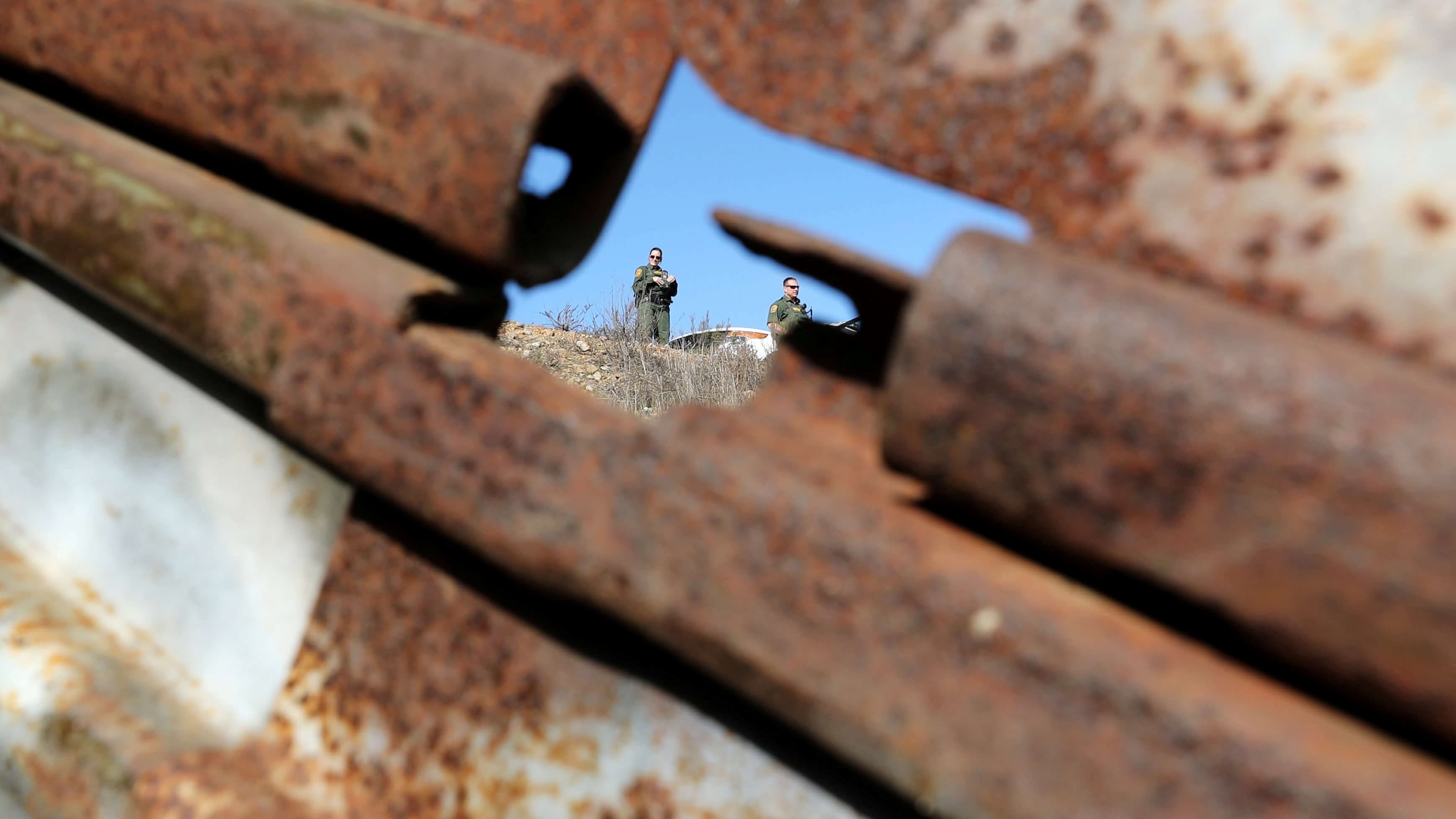 Through a hole in a metallic border wall, two Border Patrol agents visible.
