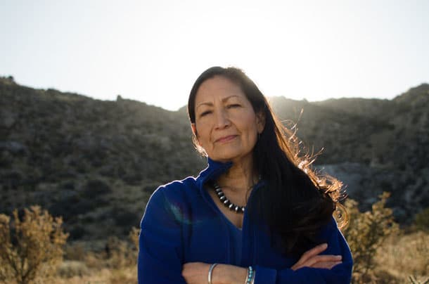 A woman with long dark hair stands outside with mountains in the background.