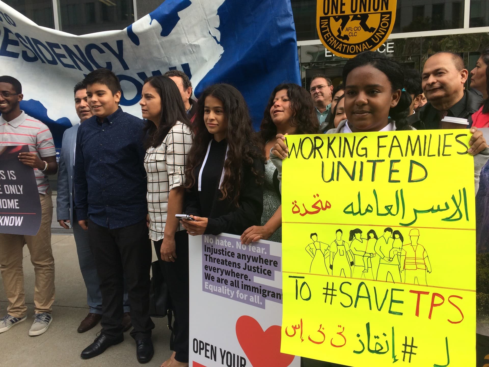 Sara Ahmed, 12, holds a sign in English and Arabic before attending a court hearing in San Francisco on Sept. 26, 2018. Cristina Morales, Crista Ramos and other TPS supporters rally nearby.