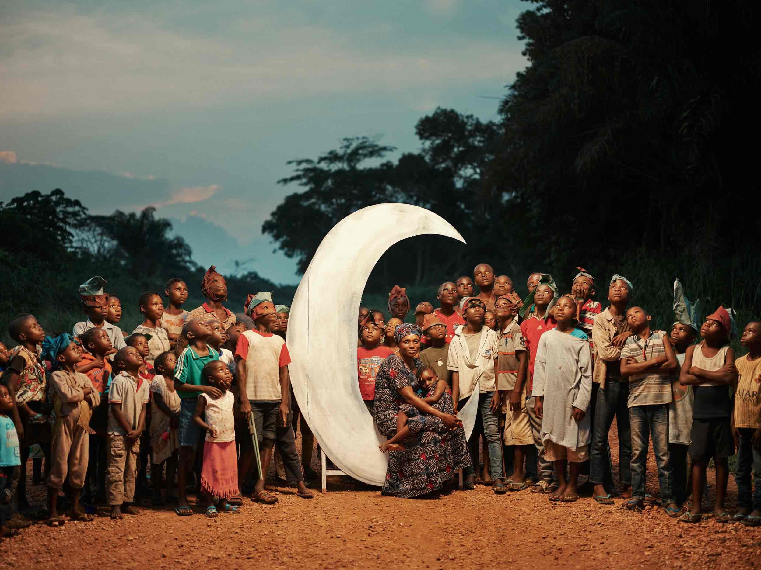 A group of children surround a woman who is seated on a large crescent moon with a child on her lap.