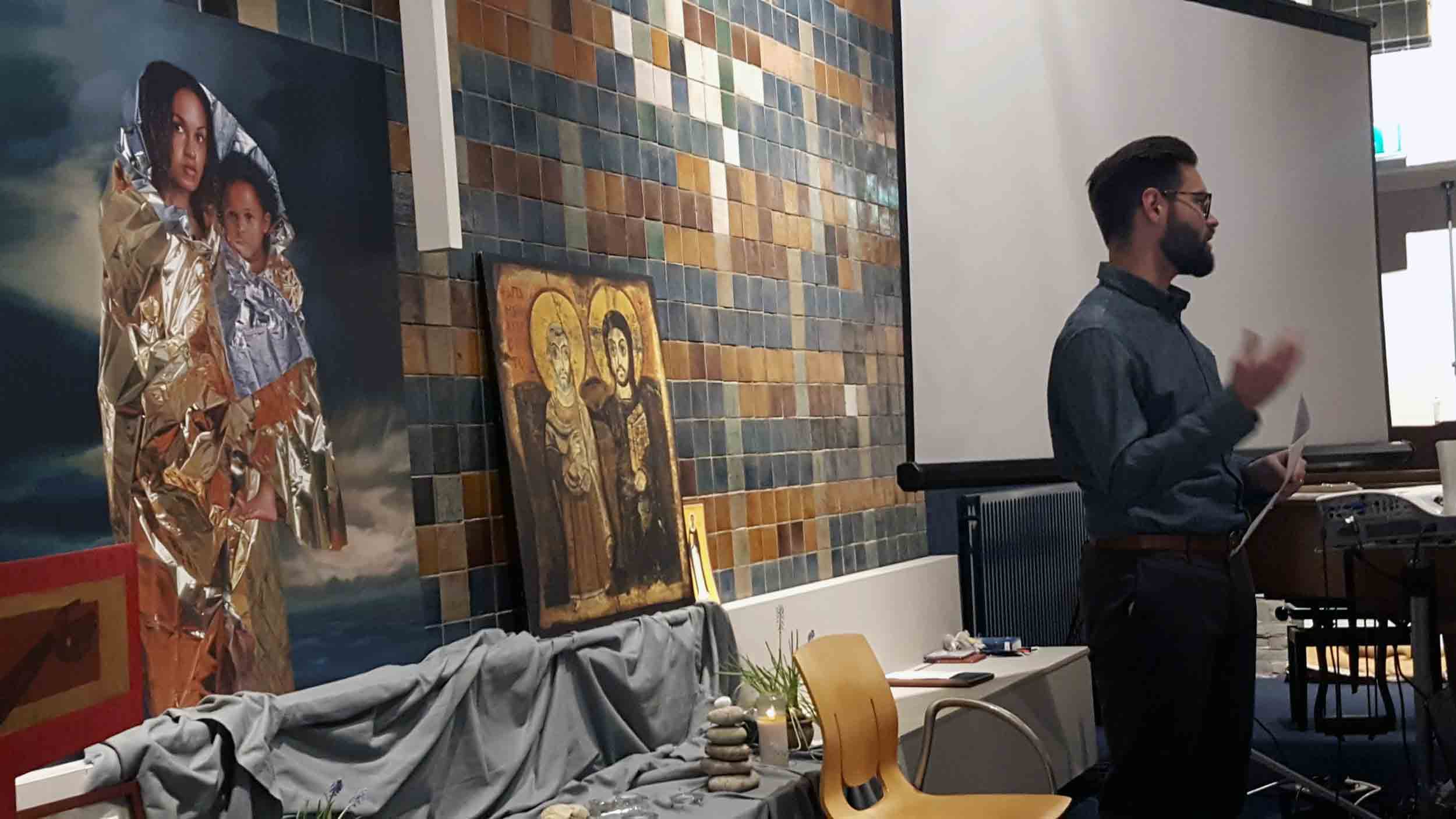 A man stands at the front of a church; behind is a picture of the Madonna with child.