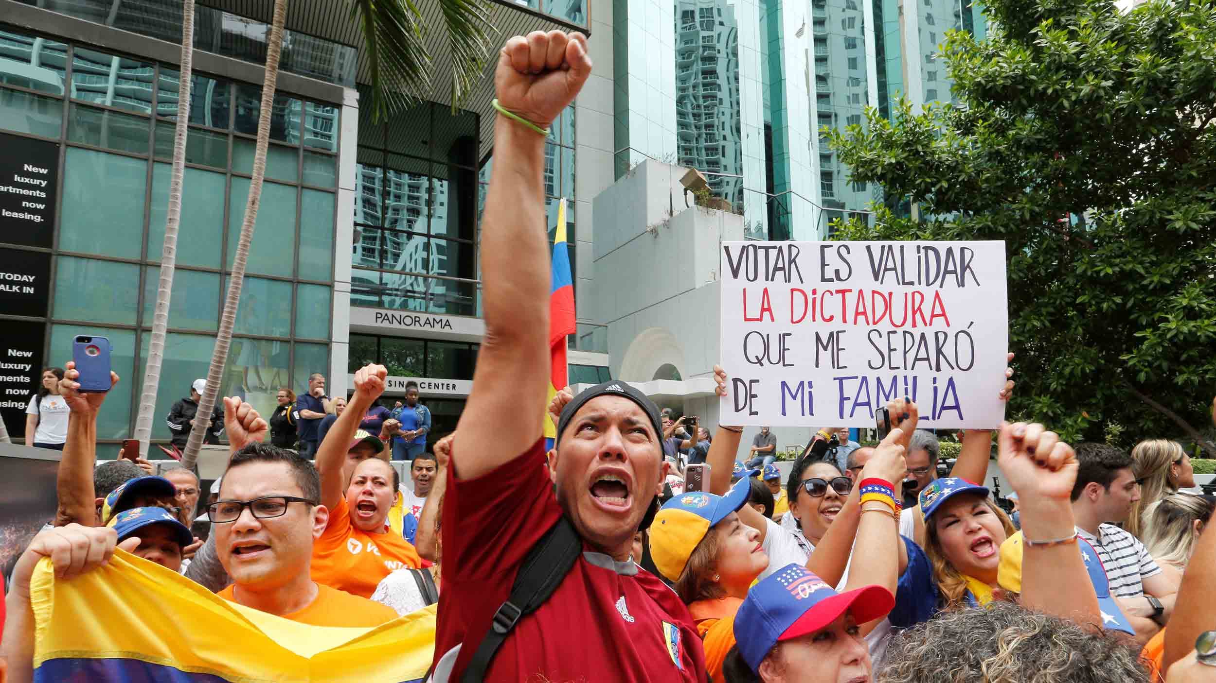 A crowd wearing yellow, blue and red of the Venezuelan flag hold signs and protest. One man in the center has his fist raised above his head.