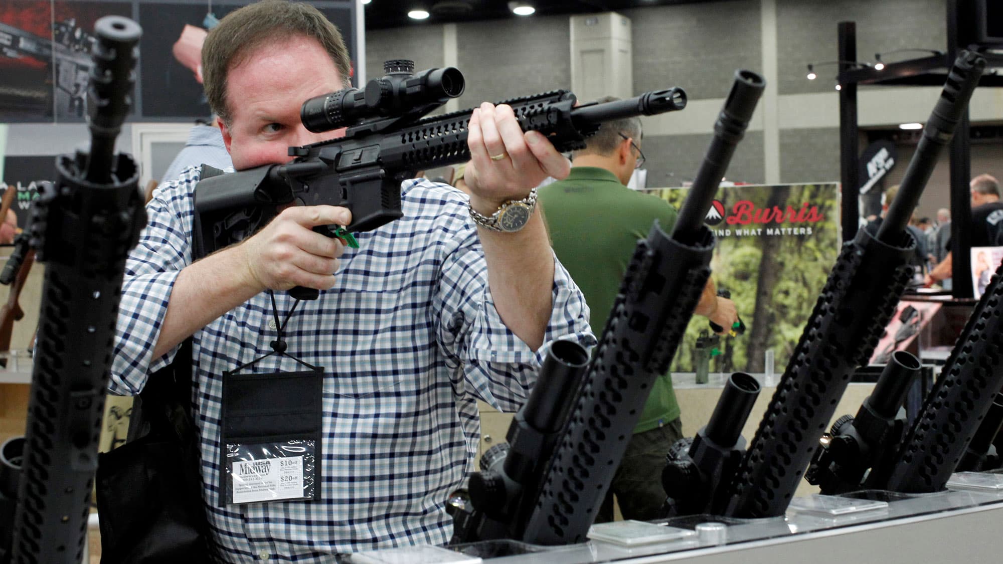 A man holds the sight of a rifle up to his right eye. More rifles are in racks on his left and right.