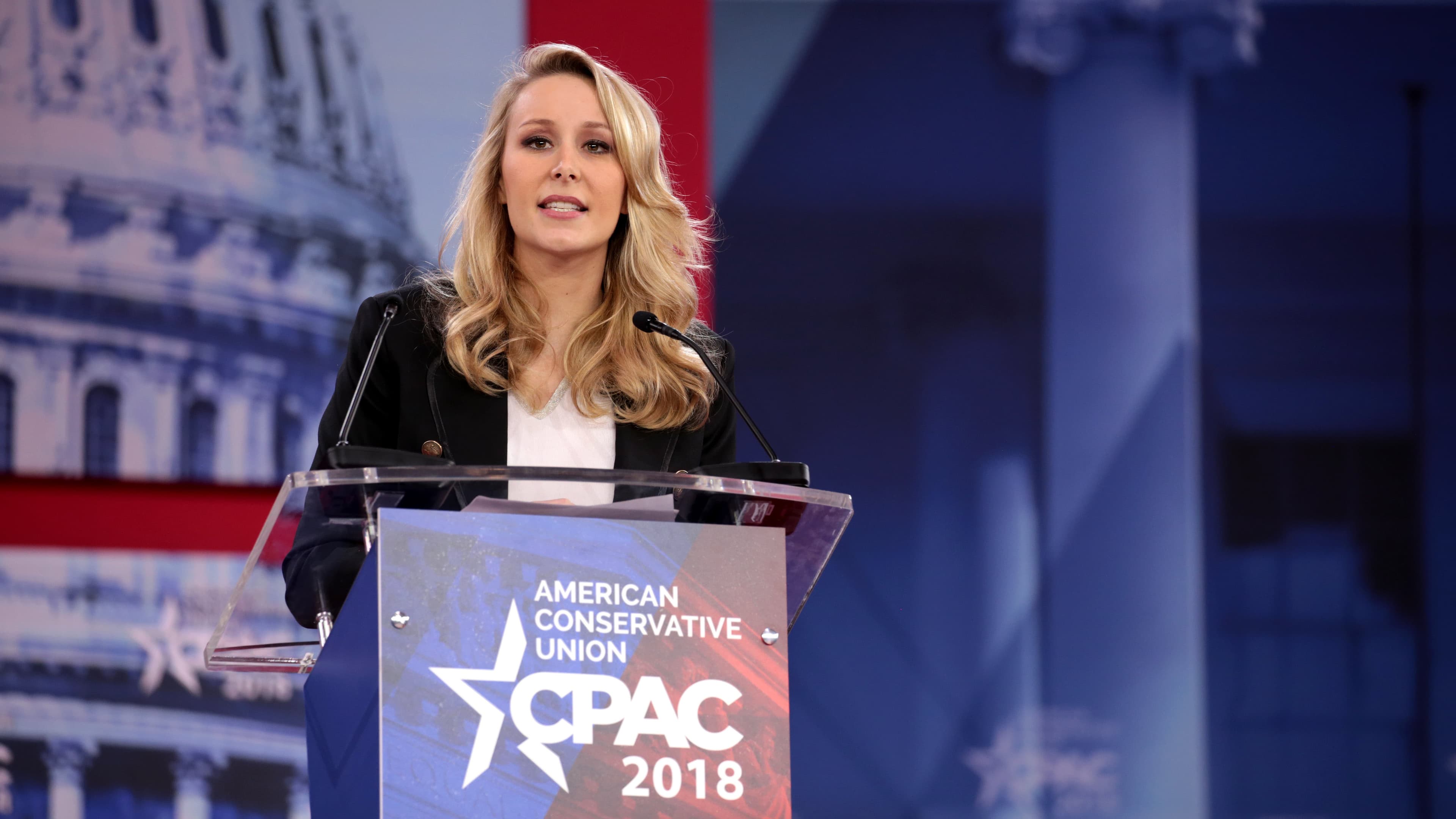 Marion Maréchal stands behind a podium and speaks in front of a backdrop of a photo of the US capitol
