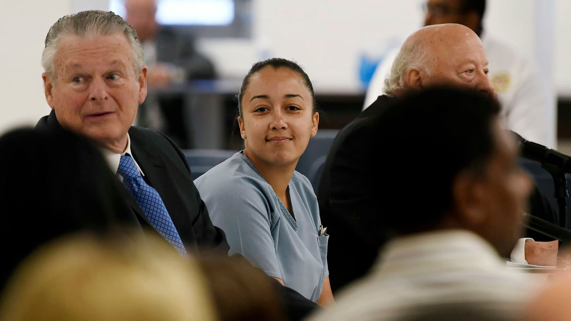 A young, light-skinned woman wearing a blue uniform sits next to two white men and smiles at family members in a court room.