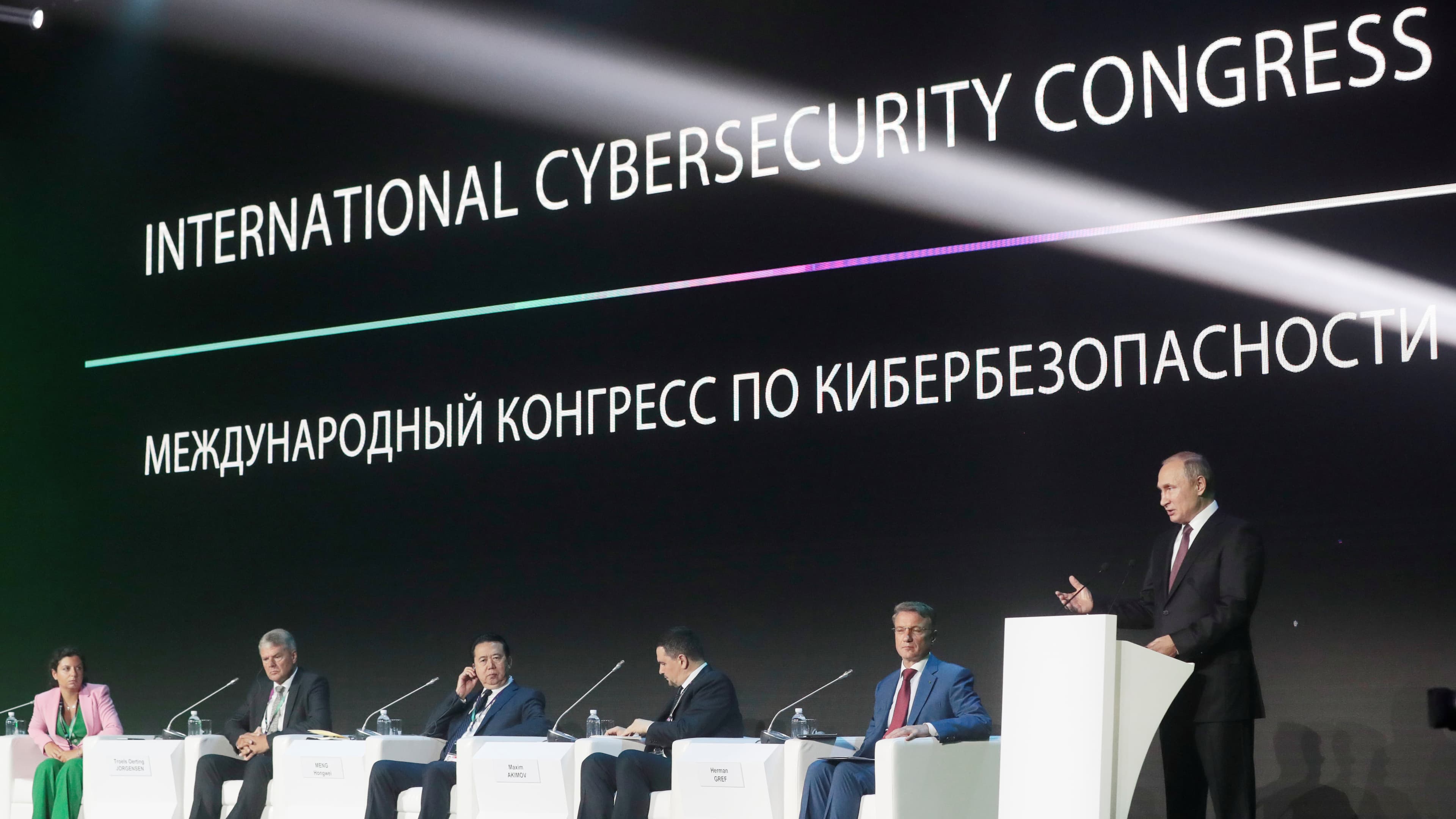 Russia's President Vladimir Putin, right, delivers a speech as 4 men and one woman sit in chairs on the stage