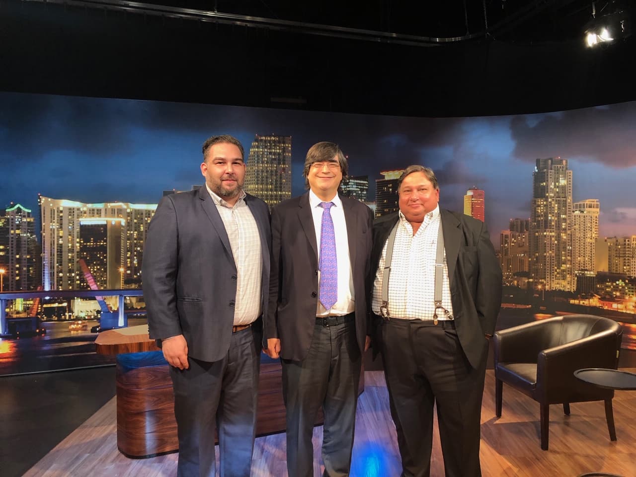 three men in suits stand on the set of a television show