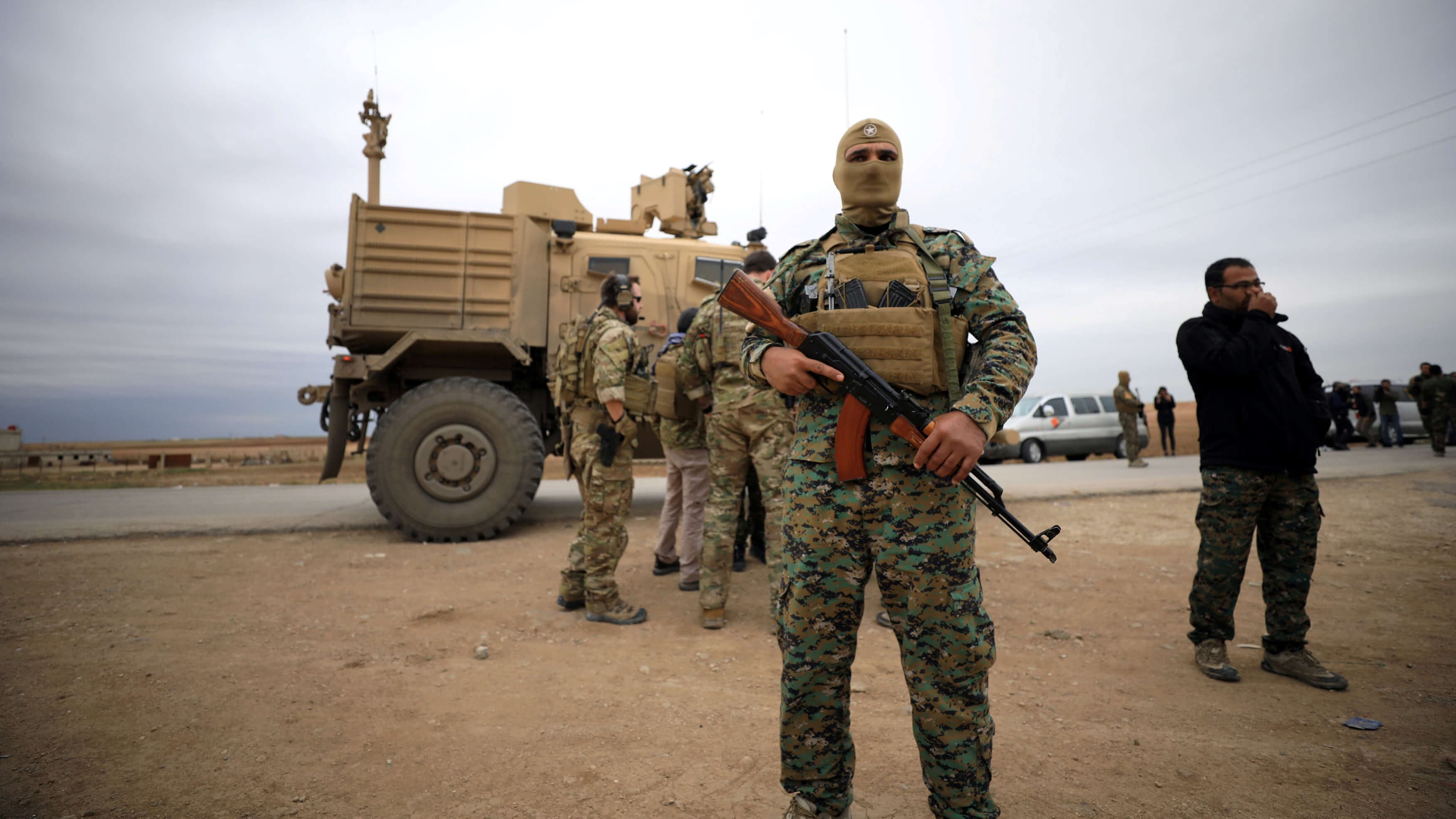 A masked Syria fighter stands in front of an American tank and some troops.