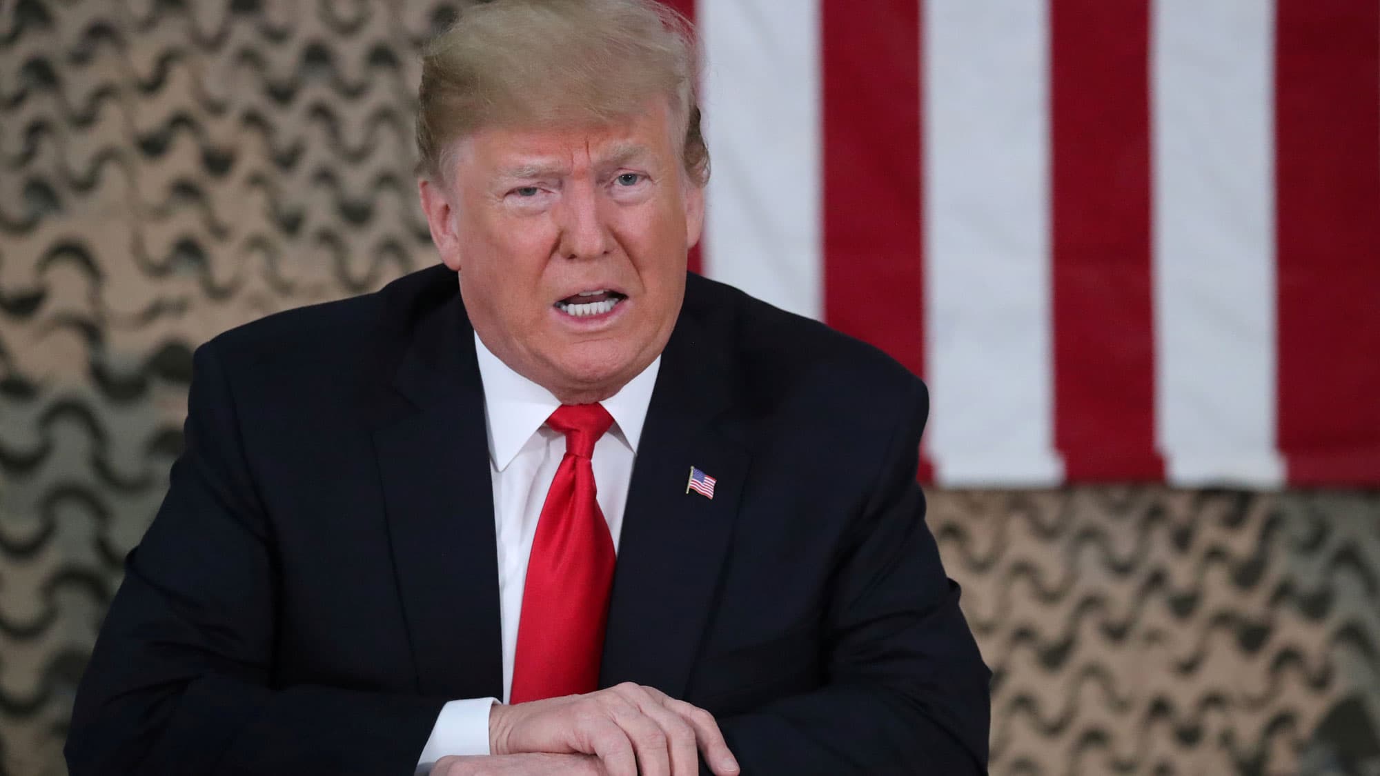 President Donald Trump speaks is shown sitting with an American flag hanging behind him while wearing a dark suit and red tie.