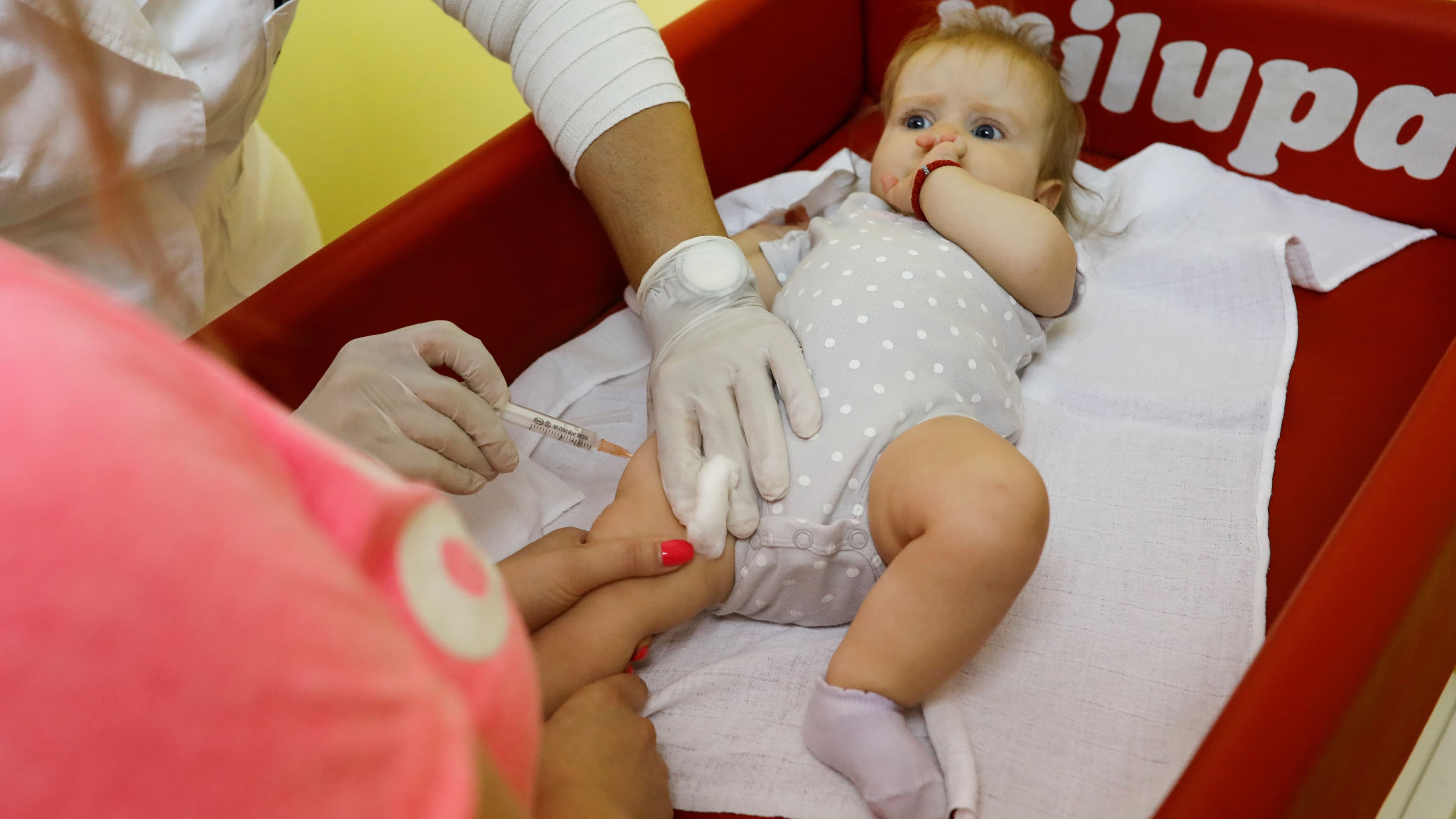 a baby with blonde hair and a white dress receives a vaccination in the left thigh