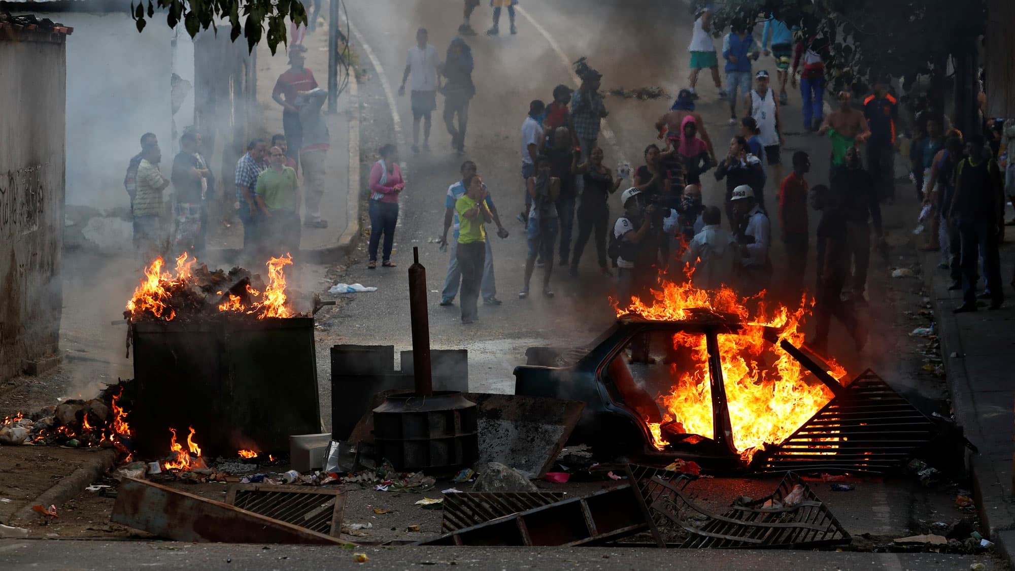 Demonstrators are shown standing behind a burning barricade comprised of a flaming car.