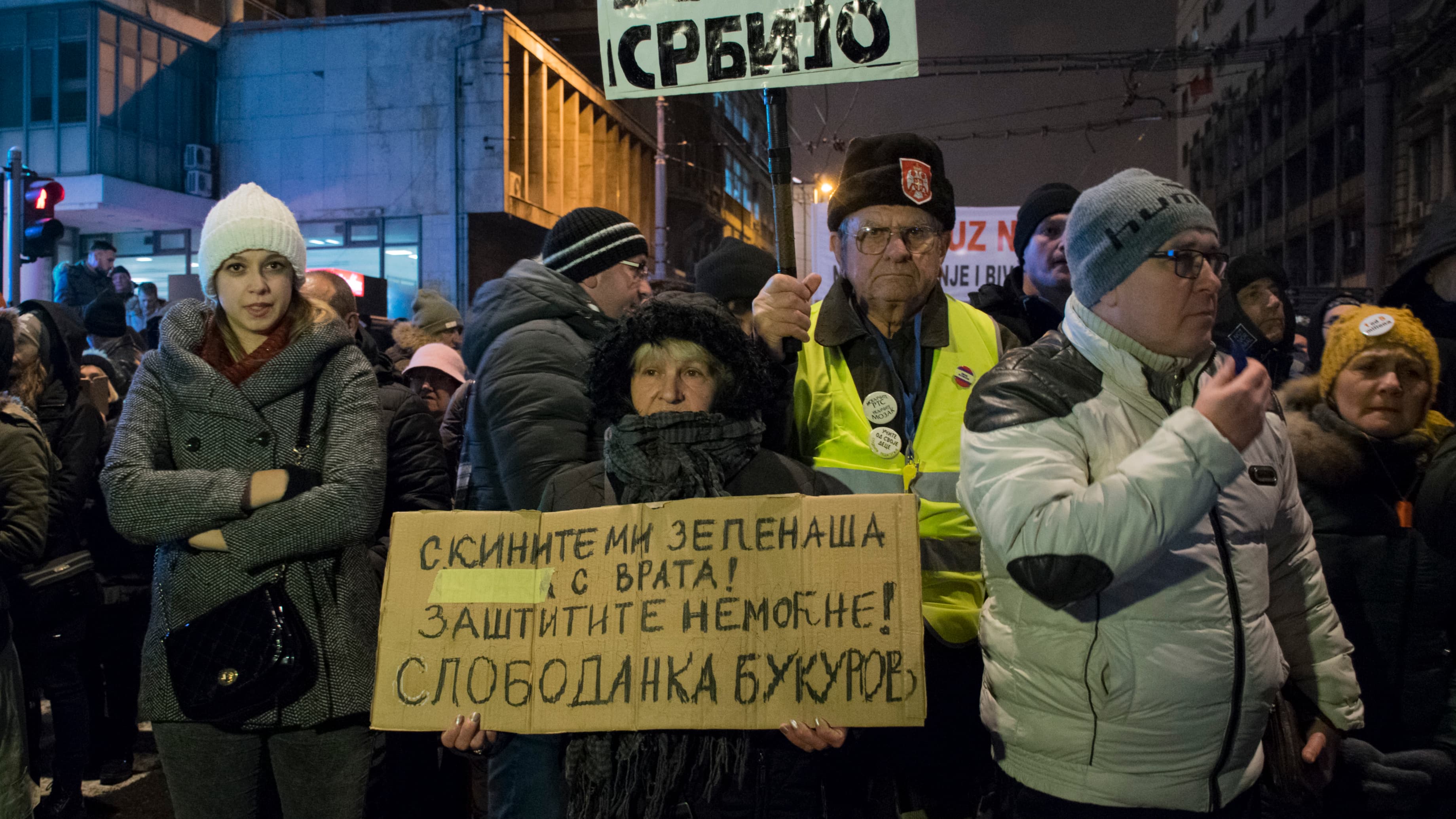 A woman stands with a cardboard protest sign, a man stands behind him with another sign.