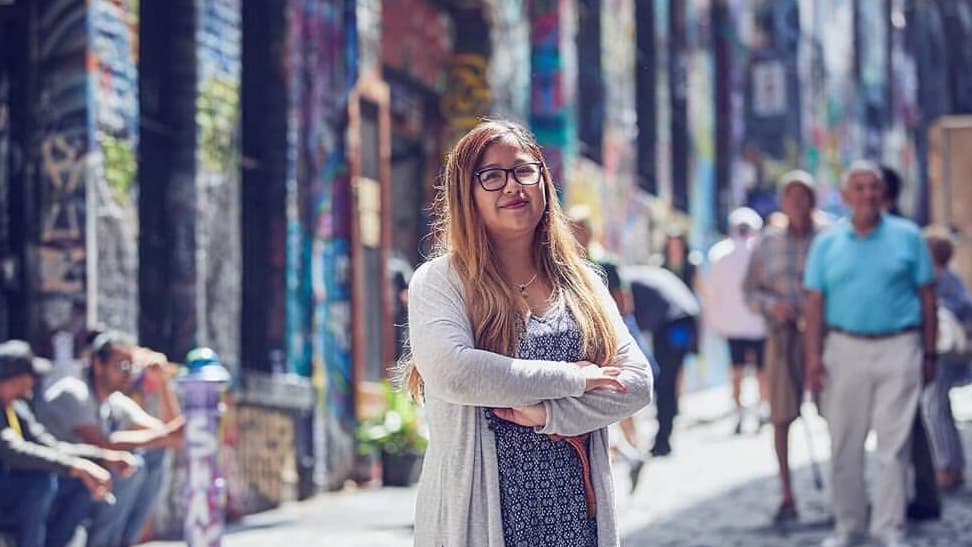 A Vietnamese Australian woman with long hair and glasses stands with her arms crossed in front of her on the street.