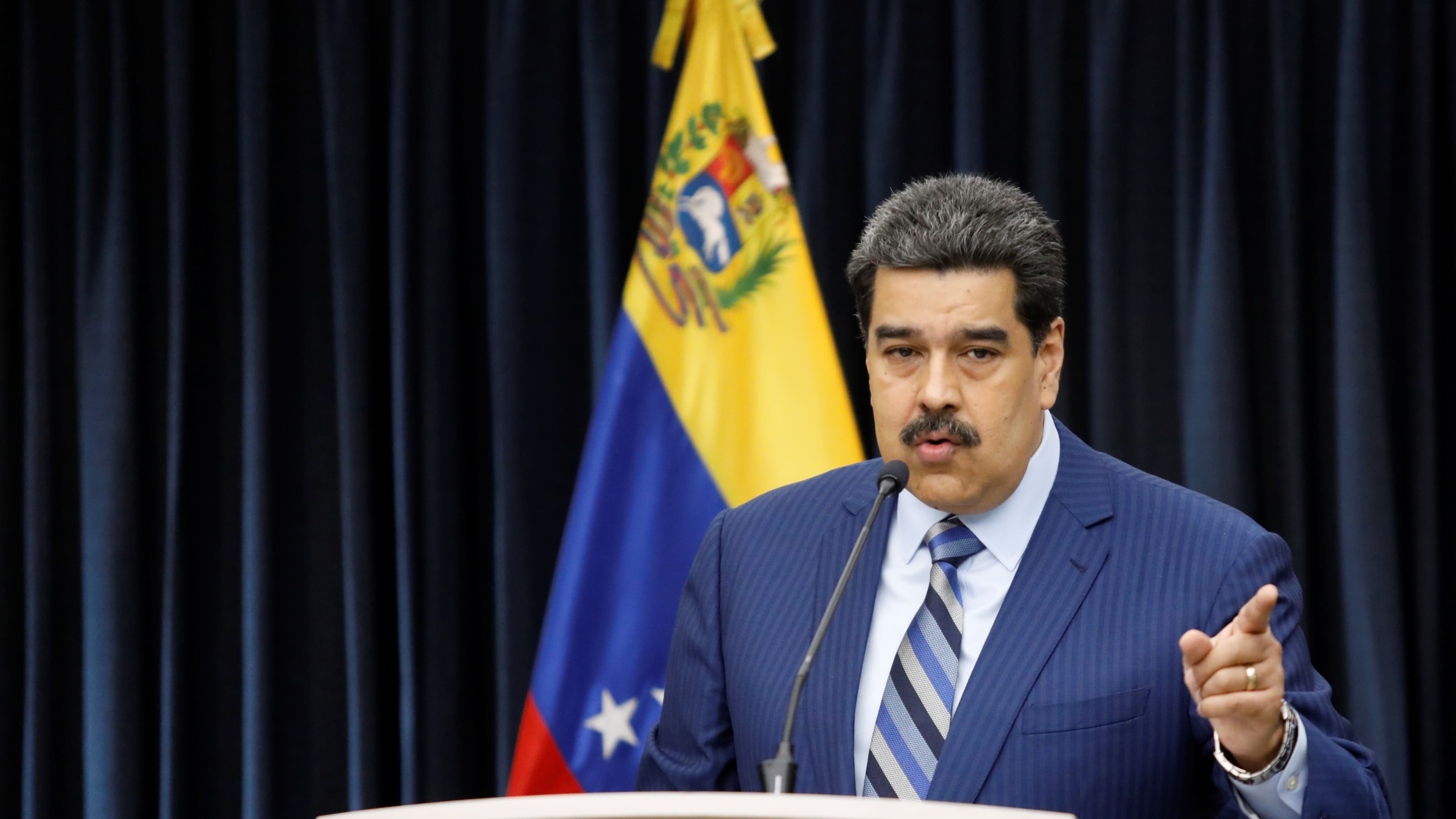 Maduro speaks at podium wearing blue suit in front of yellow and blue flag.