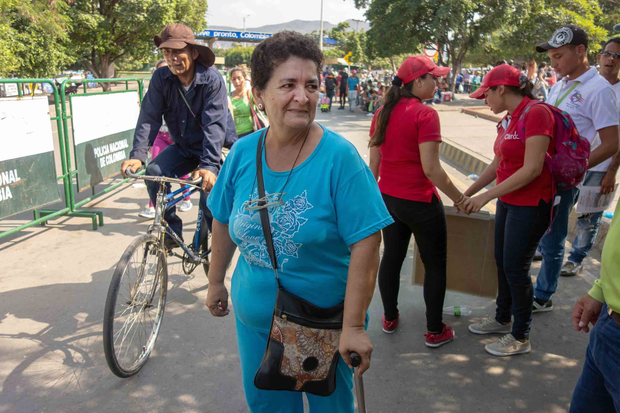 A woman in her 60s walks with a cane and wears a light blue shirt and slacks with glasses around her neck.