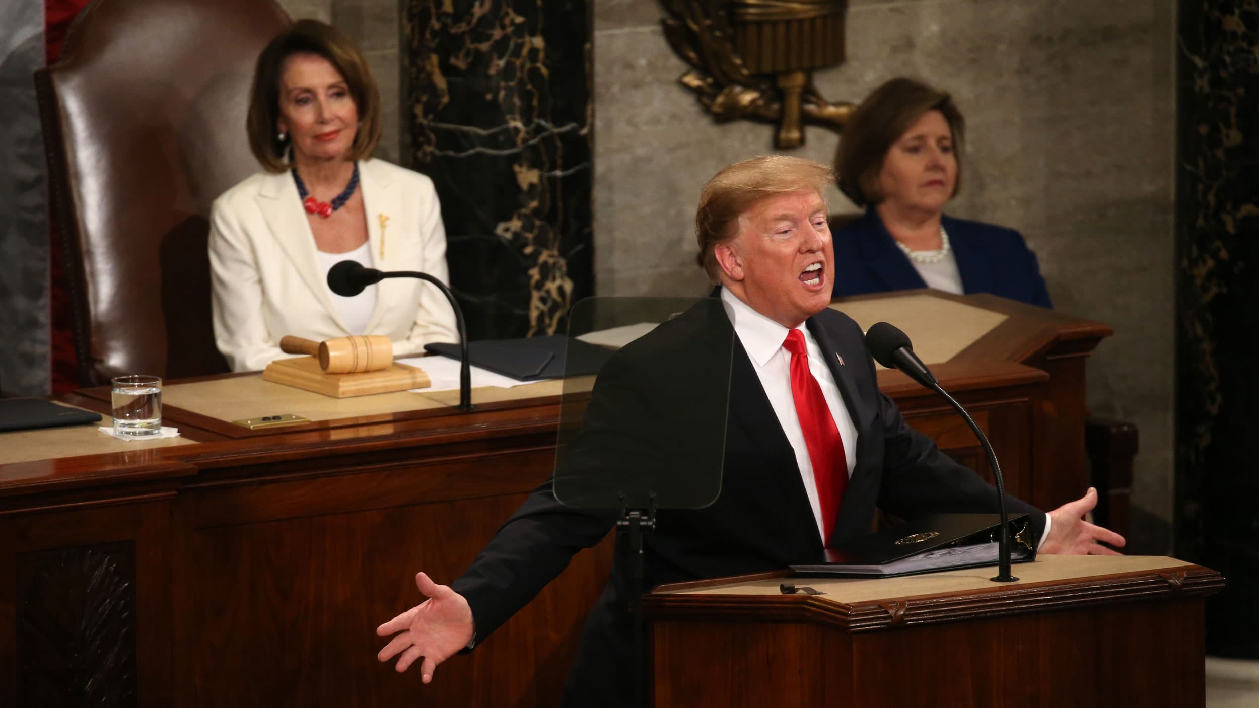 Donald Trump stands with his hands raised at the front of the House of Representatives. Behind him, Nancy Pelosi looks on.