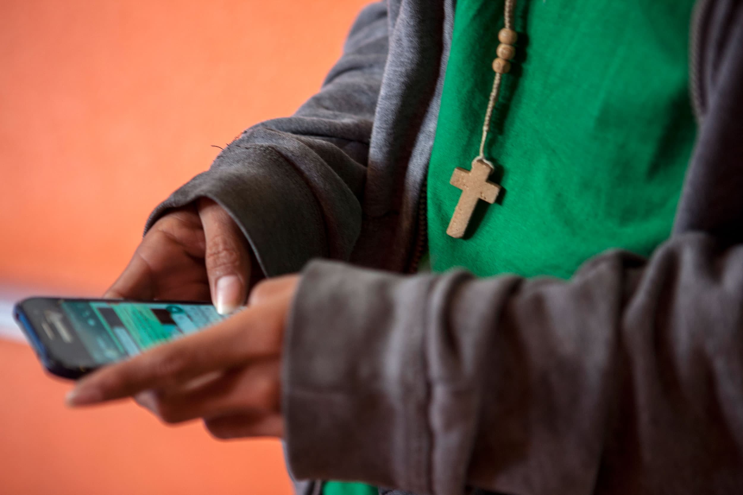 A teenage boy holds a cell phone in his hands. A wooden cross hanging from his neck dangles into the frame. His face is not pictured.